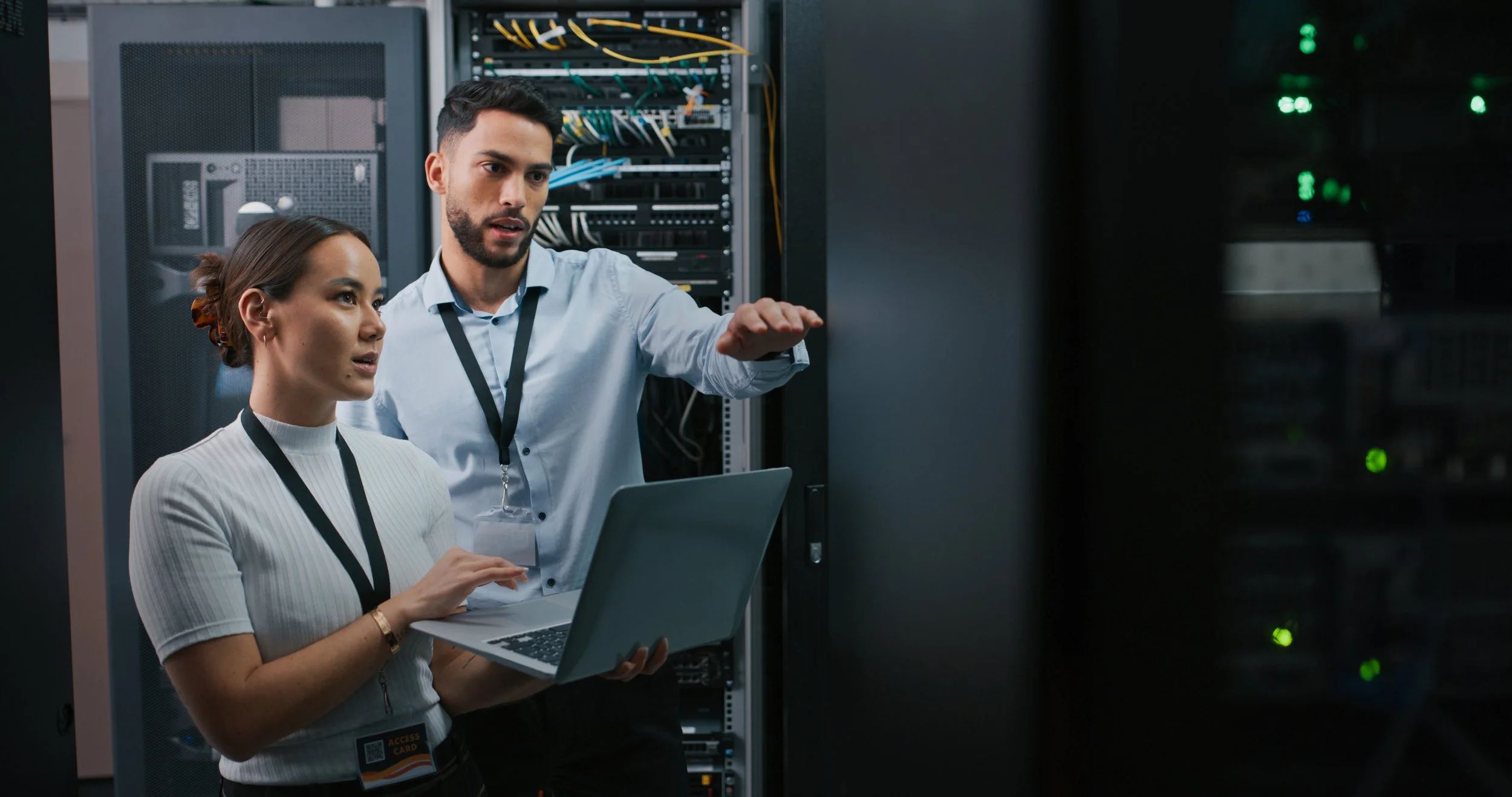 Two IT professionals working in a server room, one person operating a laptop and the other explaining something with hand gestures in front of server racks.