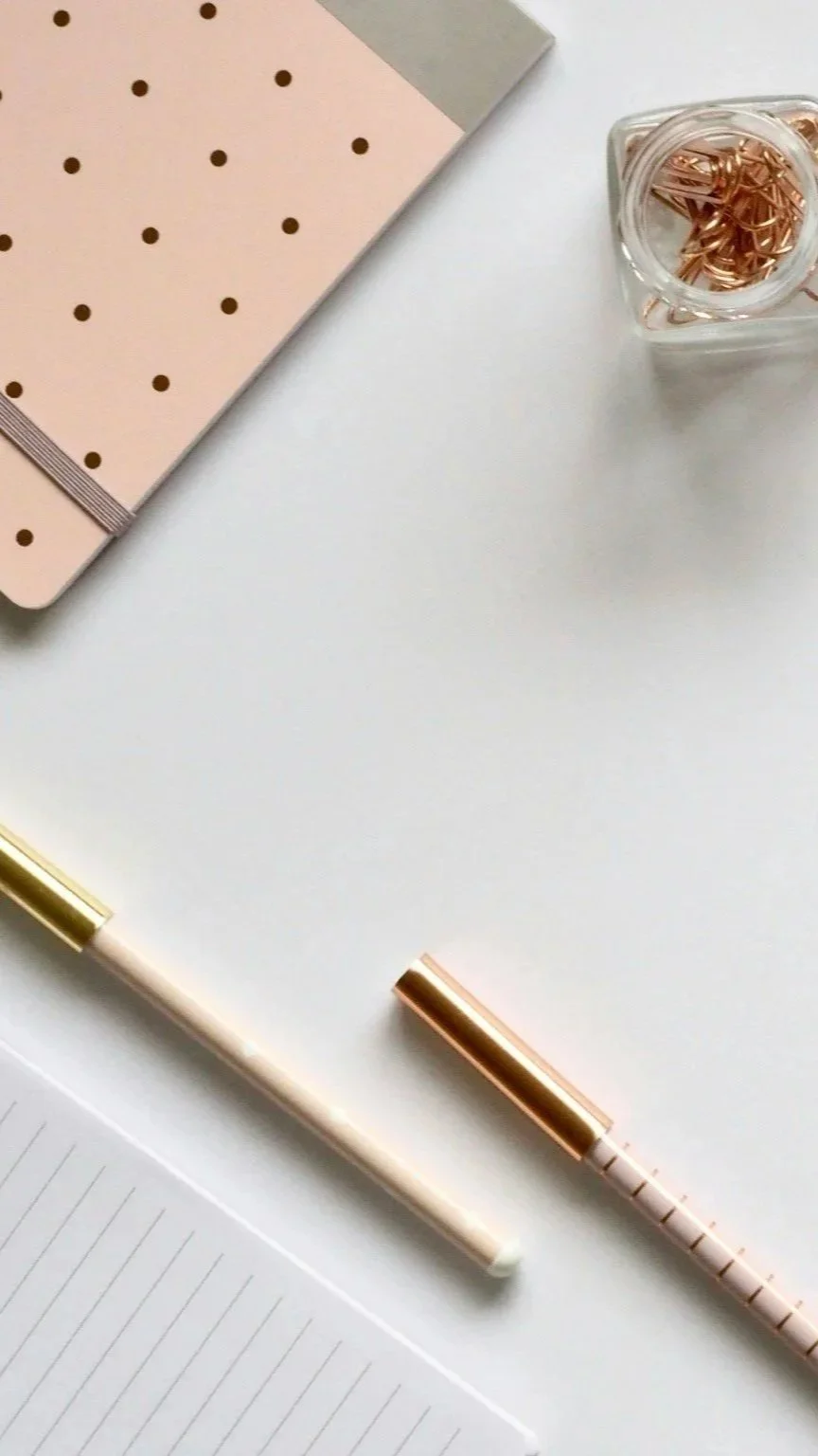 Flat lay of a white desk with a pink polka dot notebook, a gold and white pen, a lined notepad, and a clear container with gold paper clips.
