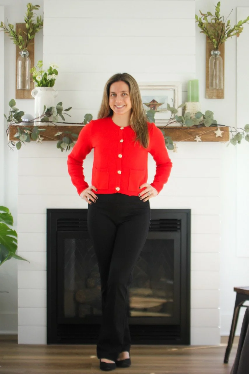 A woman standing in front of a fireplace, wearing a red cardigan with gold buttons and black pants.