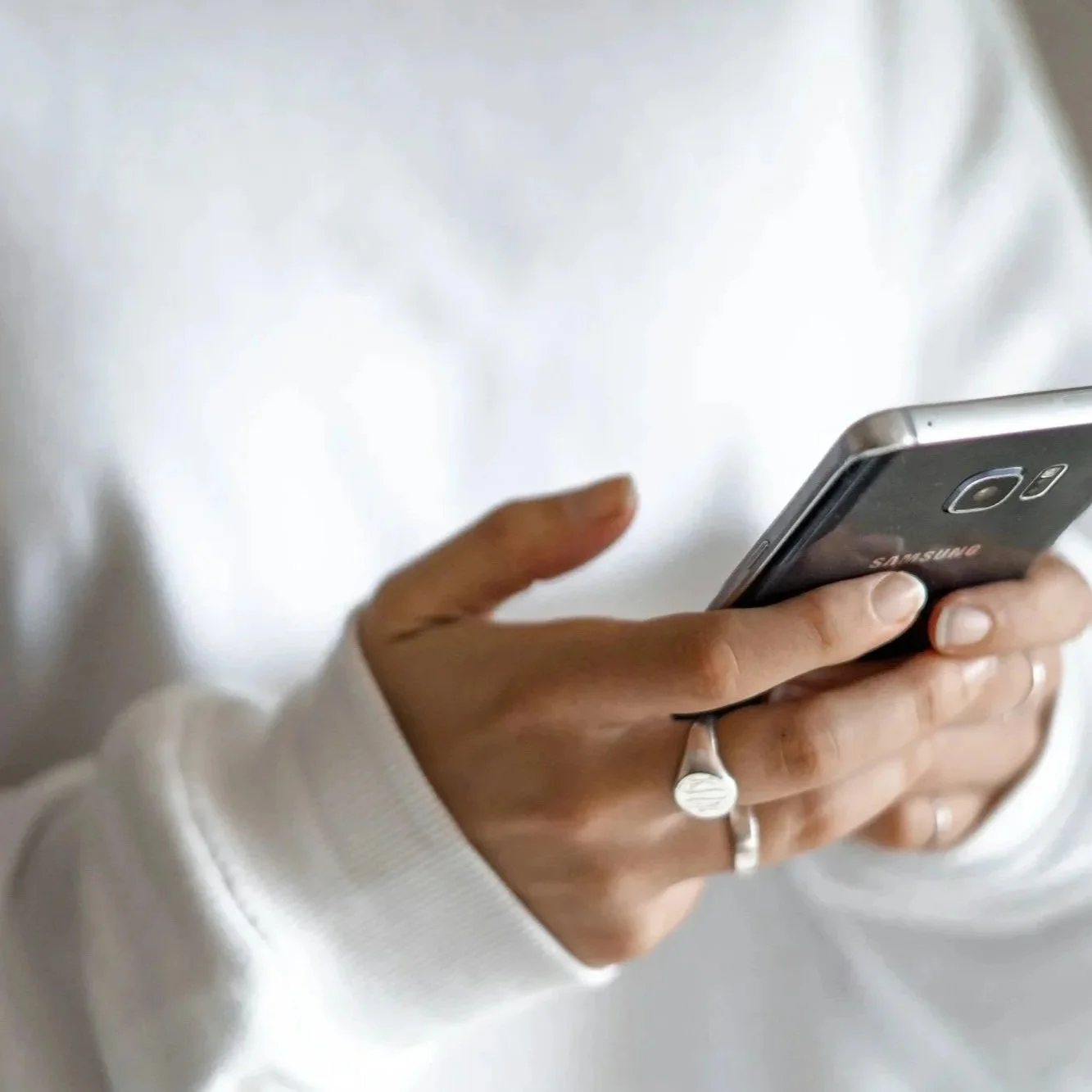 Person holding a Samsung smartphone, wearing multiple rings, including a distinctive signet ring, dressed in a white long-sleeve shirt.