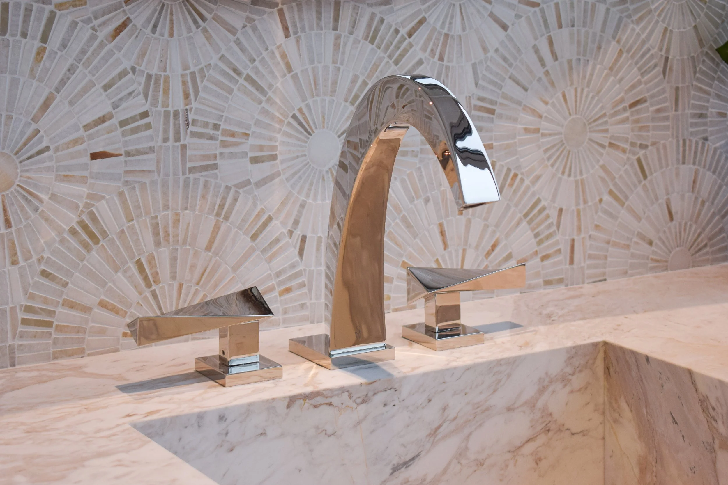 Close-up of a modern chrome kitchen faucet with separate hot and cold handles on a marble countertop, with a mosaic tile backsplash featuring a fan pattern in neutral tones.