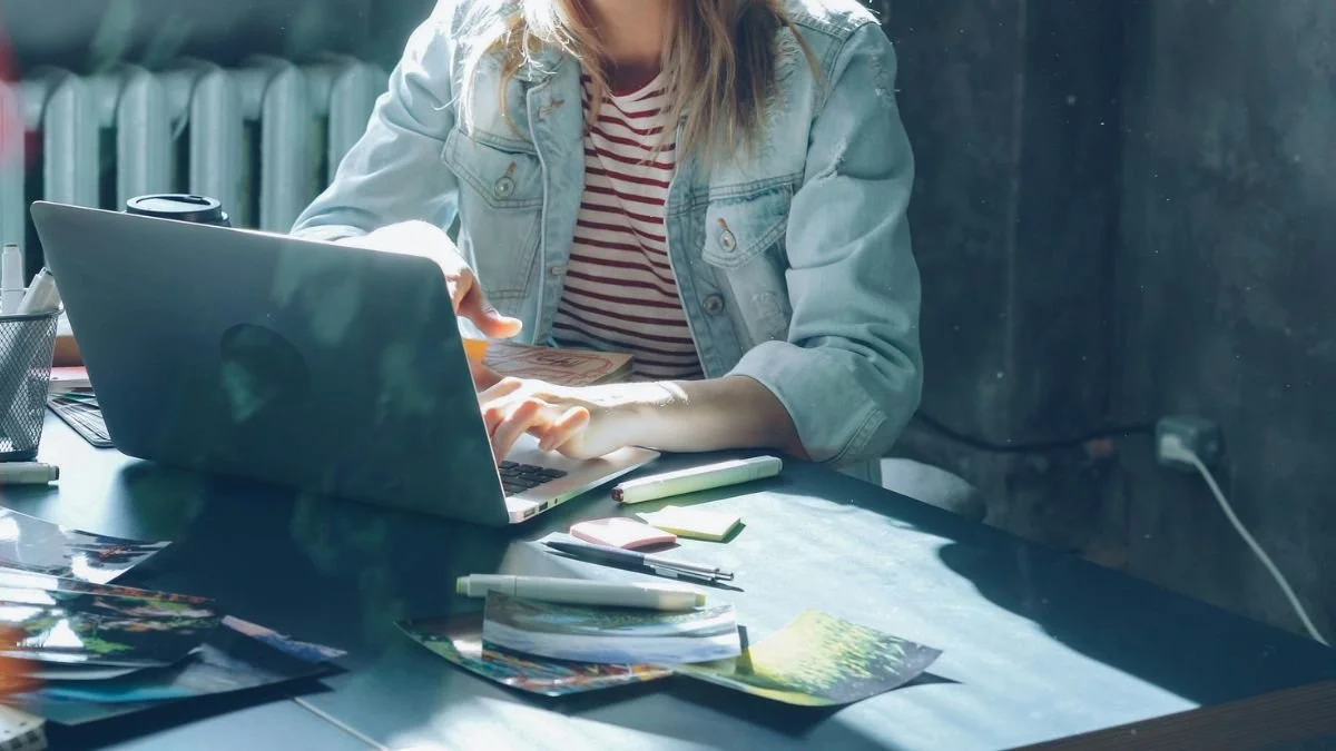 Cropped, warm shot of a woman working on a laptop, with a busy desk of pens and photos