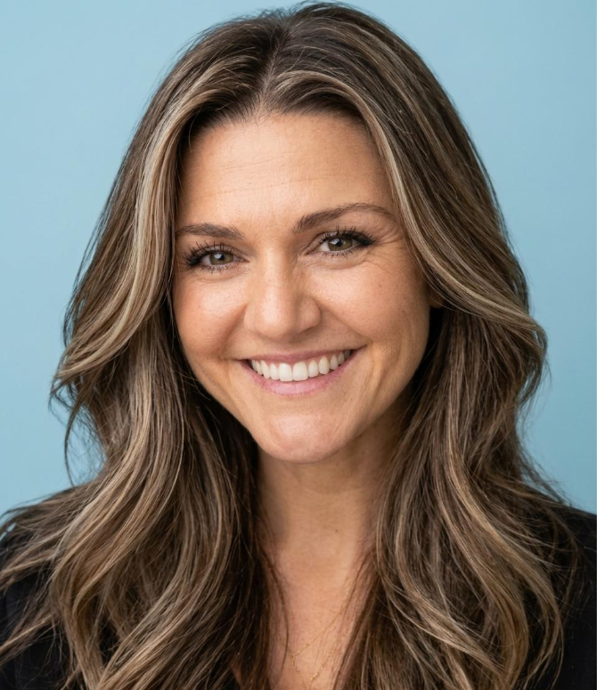 Close-up portrait of a woman with long, wavy brown hair, smiling with teeth showing, against a light blue background.