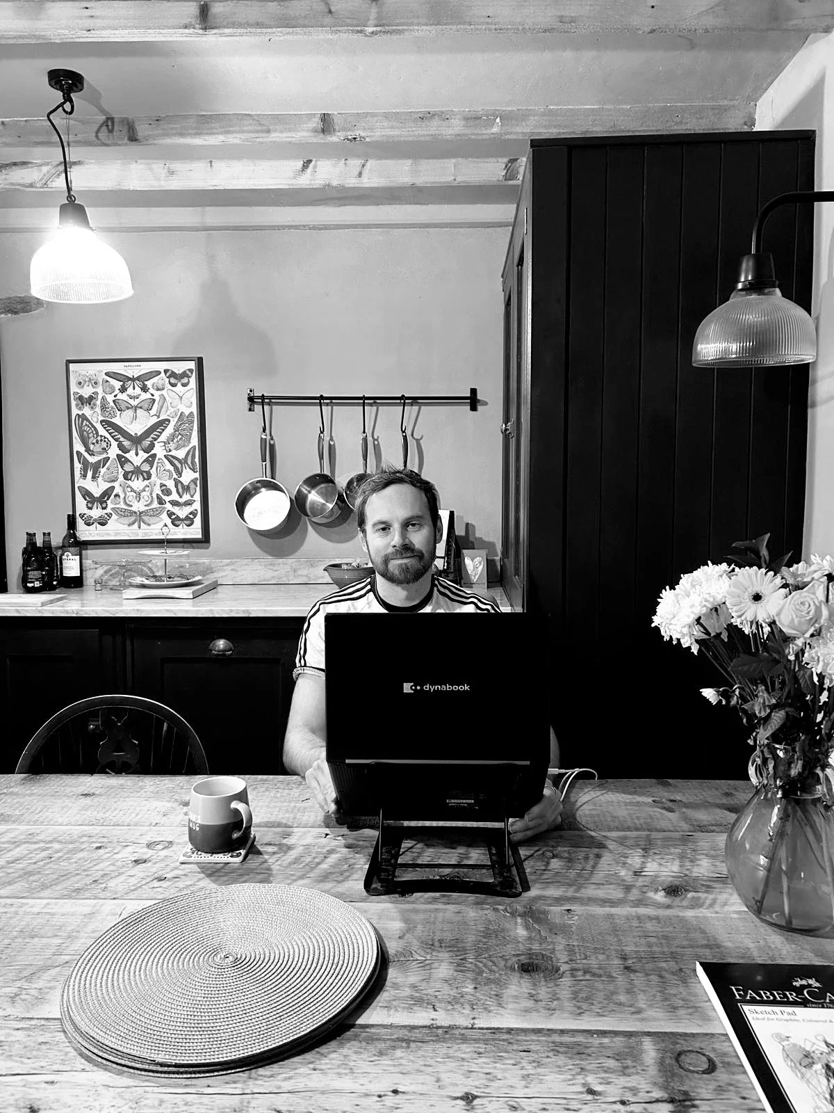 A man sitting at a wooden dining table with a laptop in front of him, in a cozy kitchen with hanging pots, a decorative poster, flowers in a vase, and some kitchen bottles.