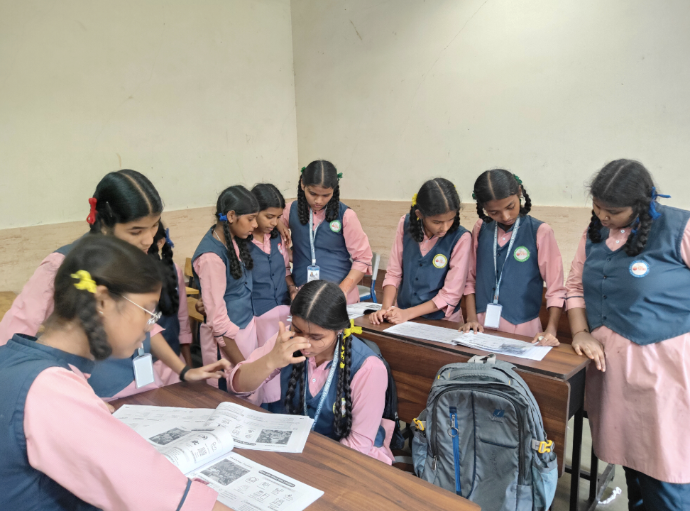A group of young girls in pink and blue school uniforms are gathered around a desk, looking at a book or magazine, with one girl in the center appearing upset or emotional. The girls are wearing ID badges and have coloring hair clips.