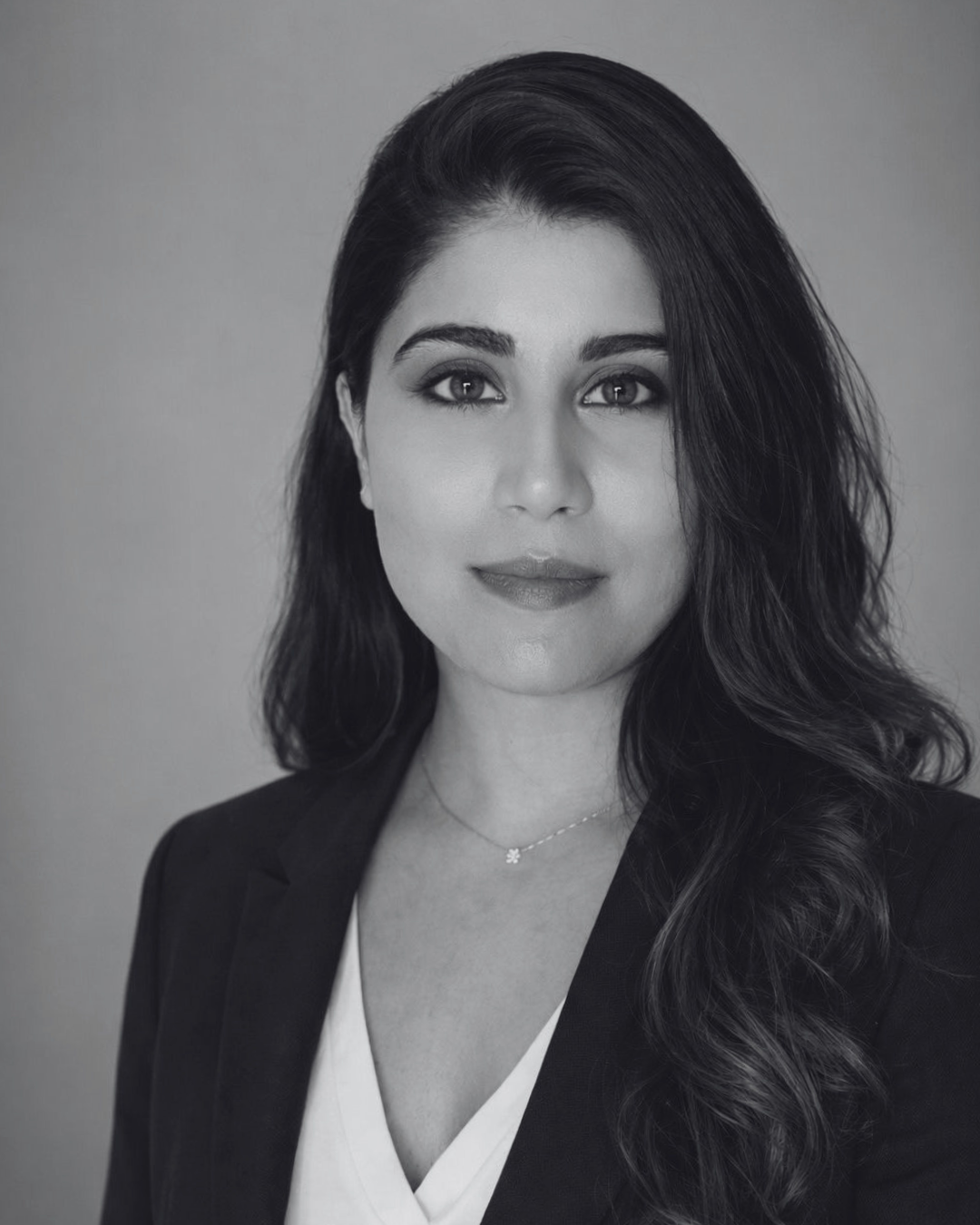 Black and white portrait of a woman with long wavy hair, wearing a blazer over a V-neck top, and a delicate necklace.