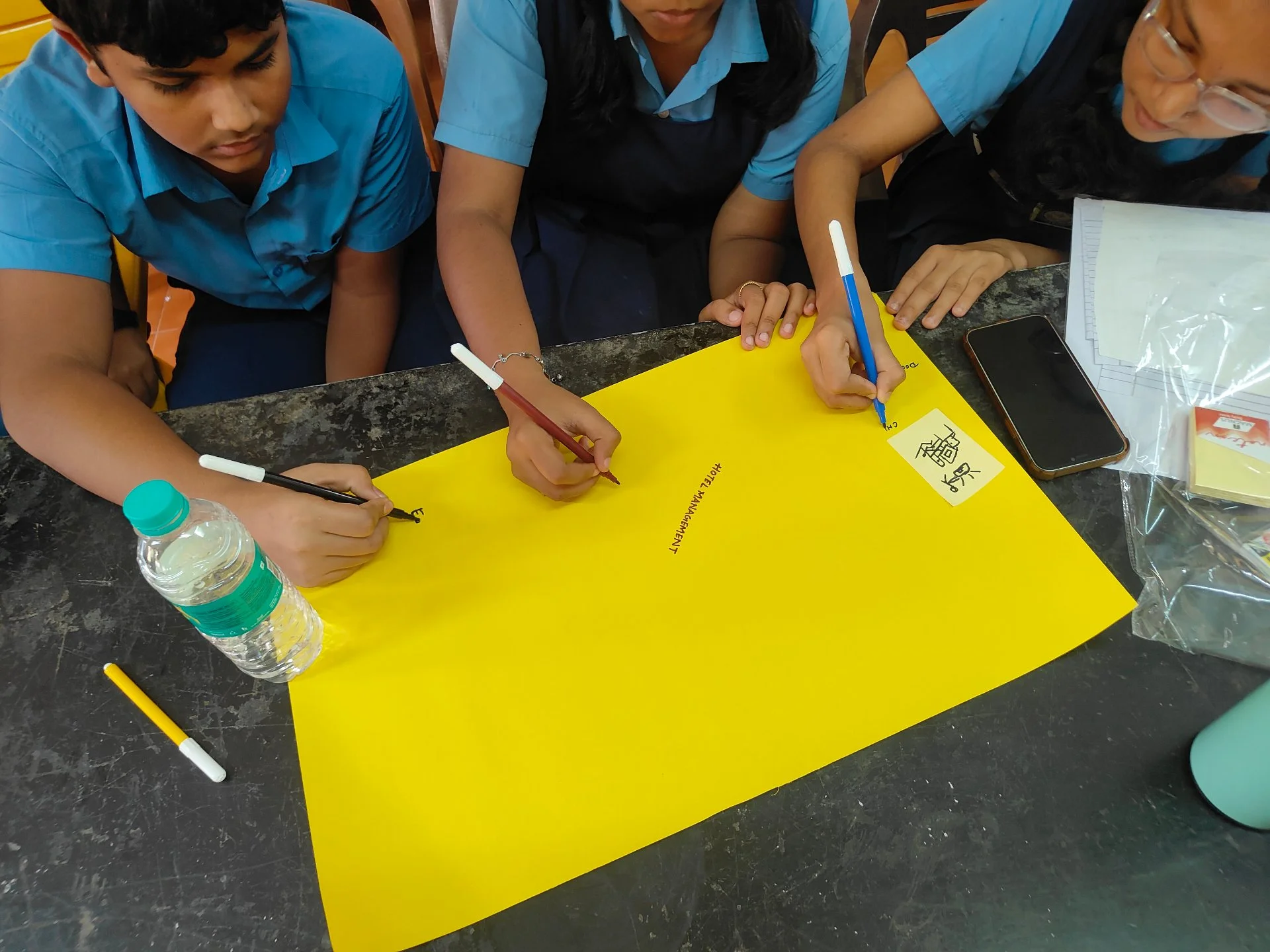 Three students are working together on a project at a table. They are writing on a large yellow poster with markers. One student is writing a word, and there is a smartphone, a water bottle, and some papers on the table.