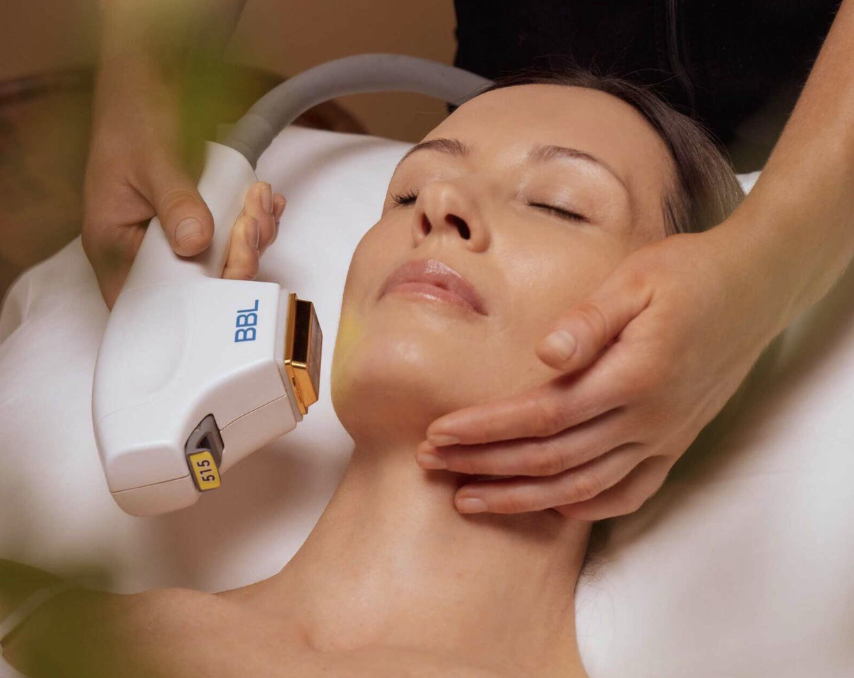 A woman is receiving a cosmetic or skincare treatment with a handheld device on her face and neck while lying down with her eyes closed.