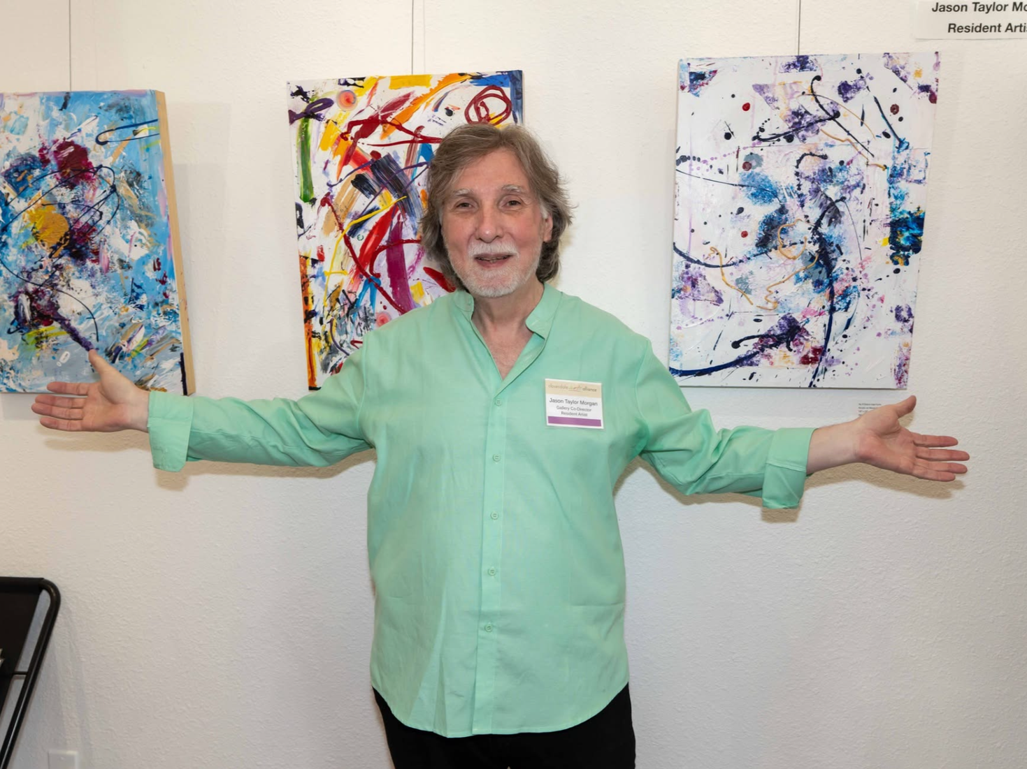 A man with gray hair and beard wearing a light green shirt stands with arms outstretched in front of three colorful abstract paintings on a white wall. He is smiling and has a name tag.
