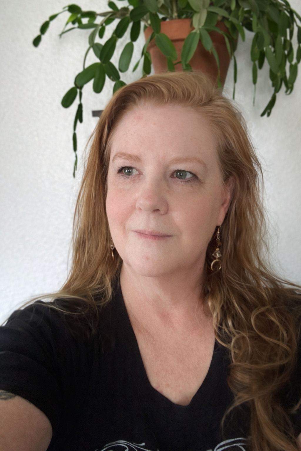 Close-up of a woman with long, curly red hair, wearing earrings, in front of a white wall and a potted green plant.