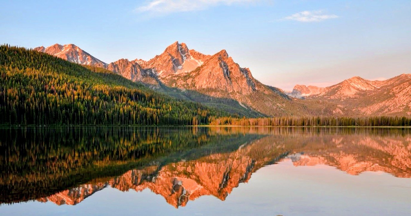 A mountain landscape with snow-capped peaks reflected in a calm lake surrounded by a forest