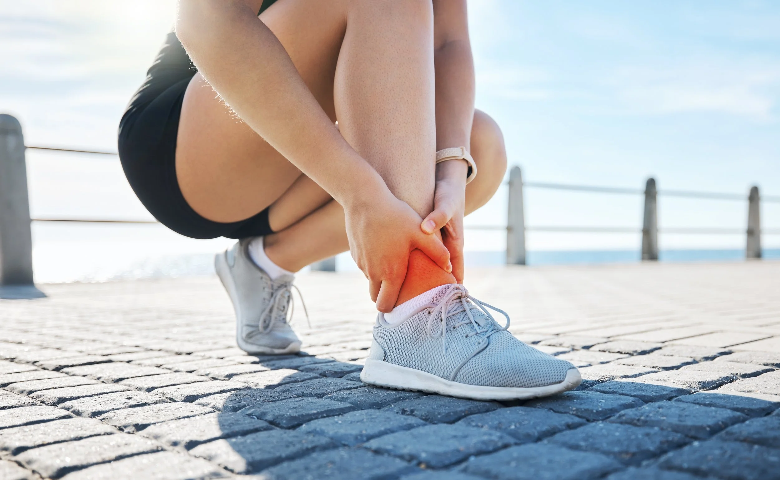 A person crouching on a paved path by the water, clutching their ankle in pain, with a bright outdoor setting and railing in the background.