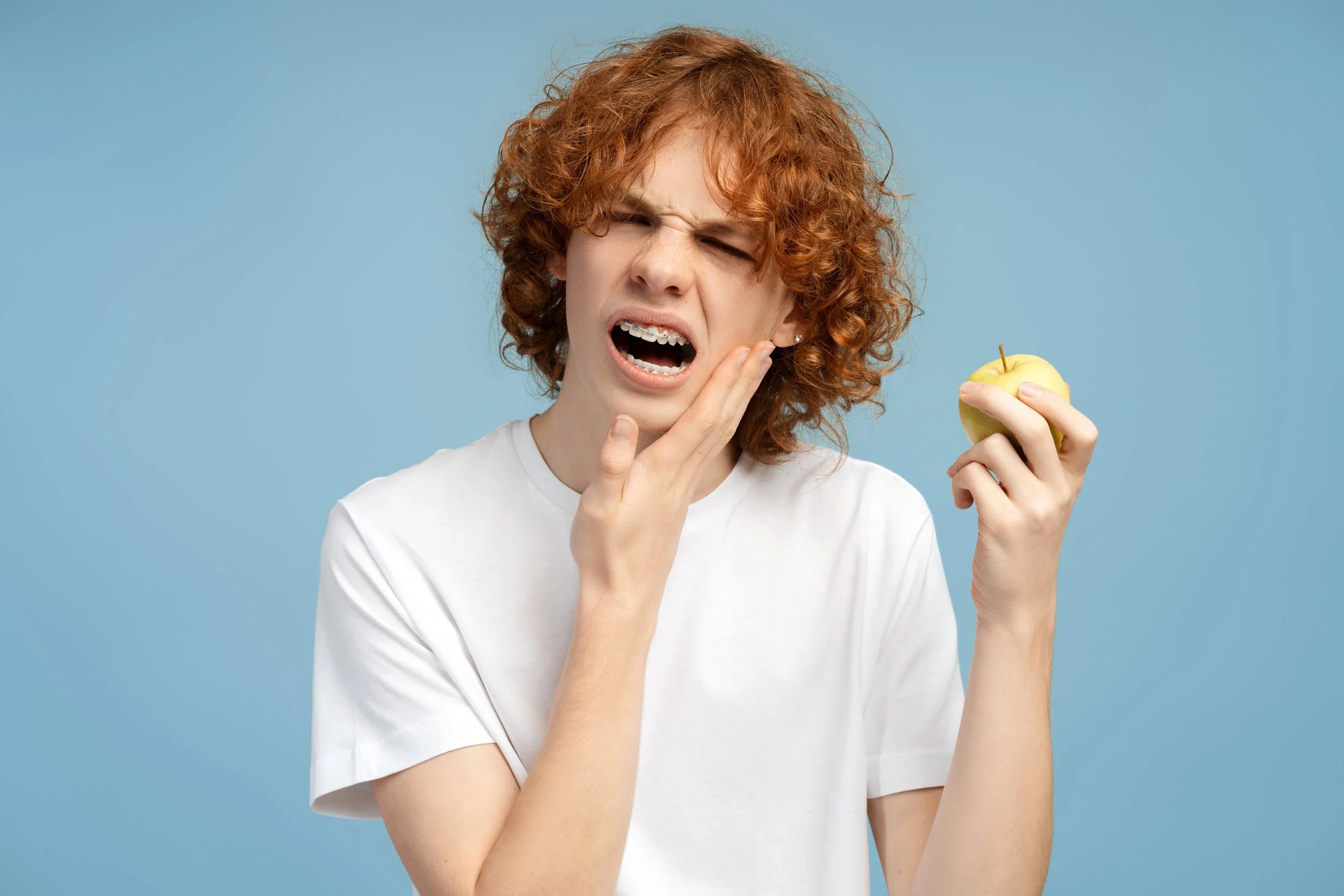 A young woman with curly red hair wearing a white t-shirt, holding a yellow apple, showing an expression of discomfort or pain, touching her cheek.