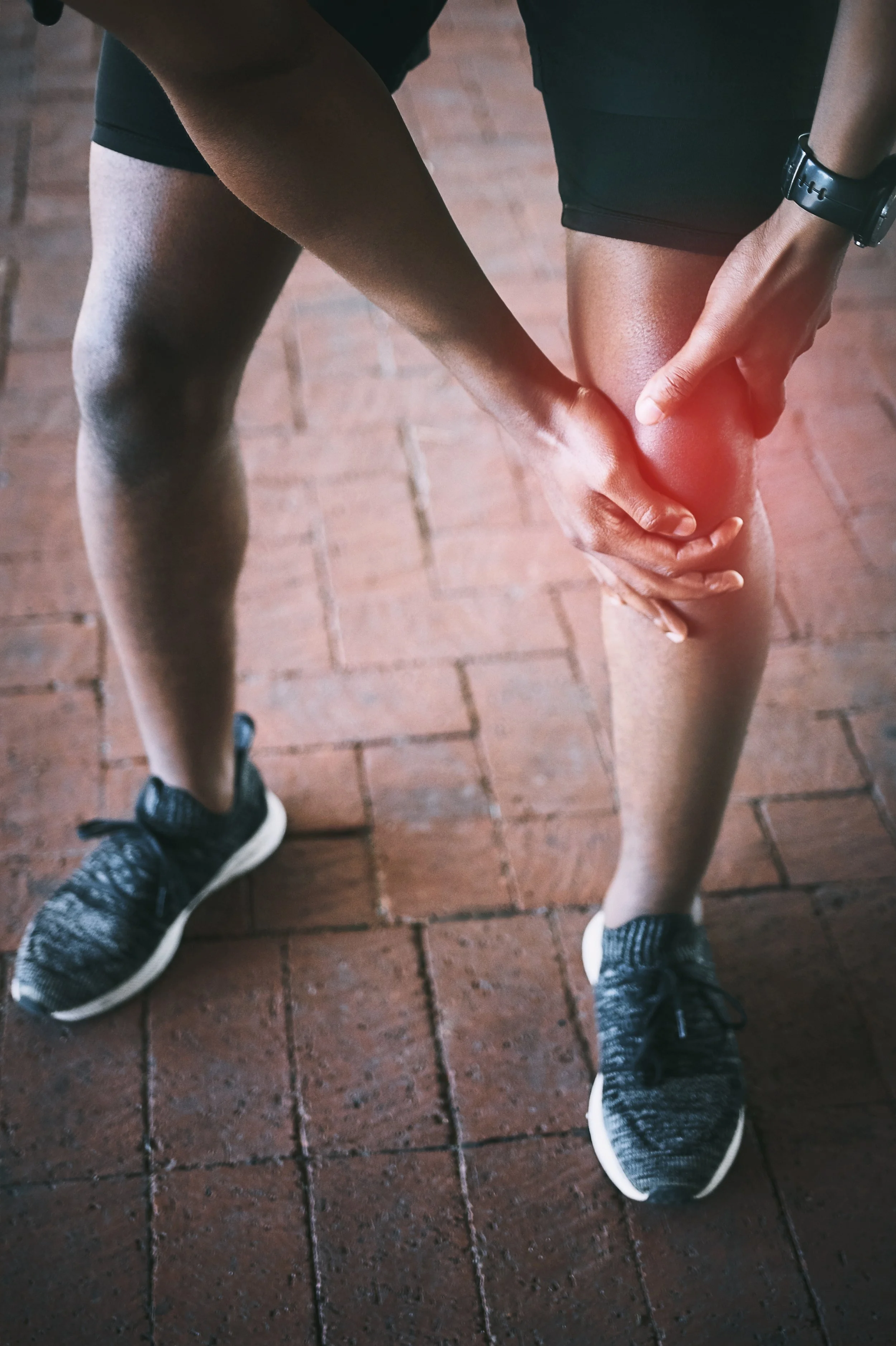 Person holding their injured knee, which is red and glowing, while standing on brick pavement. The person is wearing black shorts, a wristwatch, and black running shoes.