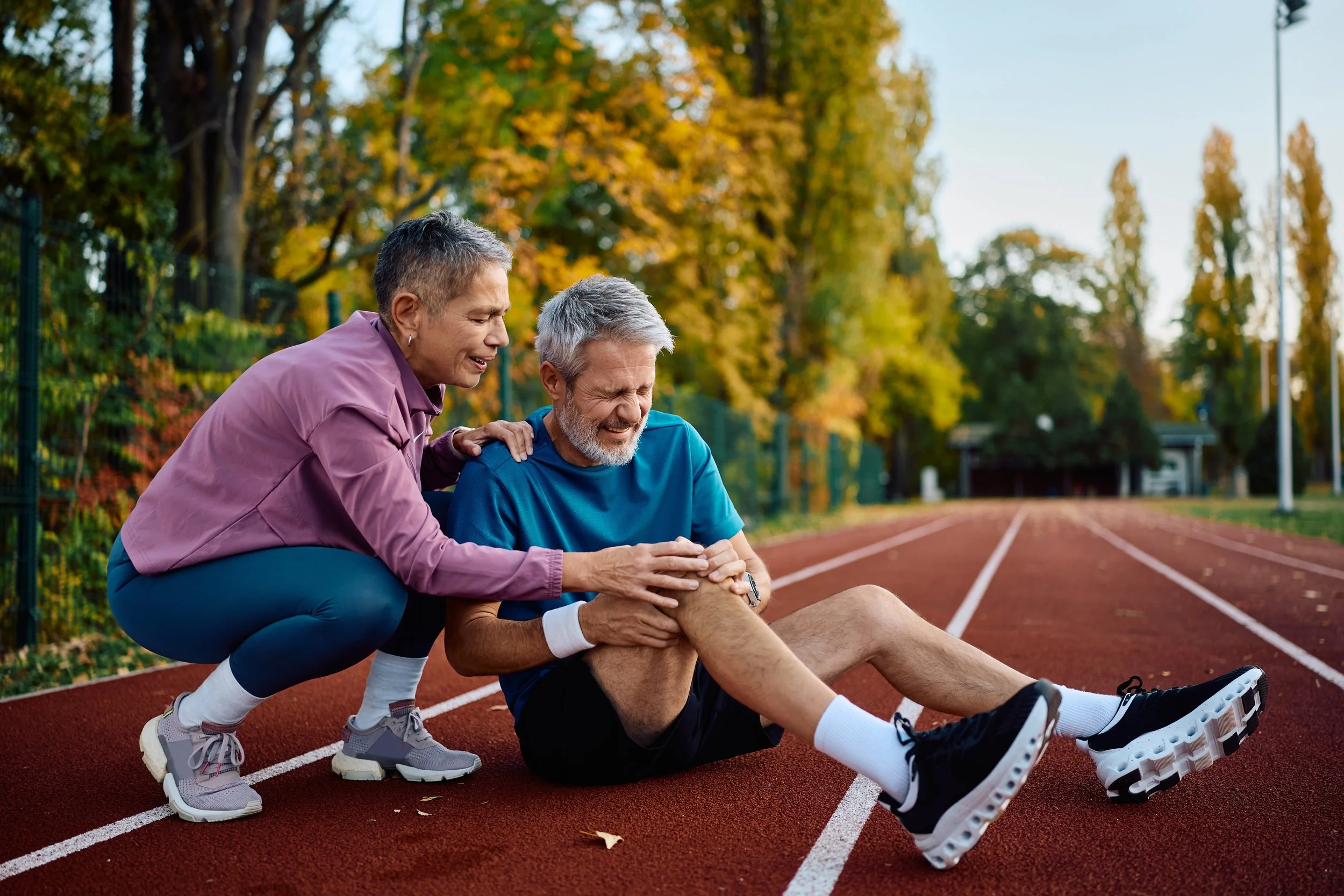 An elderly man appears to have hurt his knee and is sitting on a running track, receiving help from a woman during outdoor exercise.