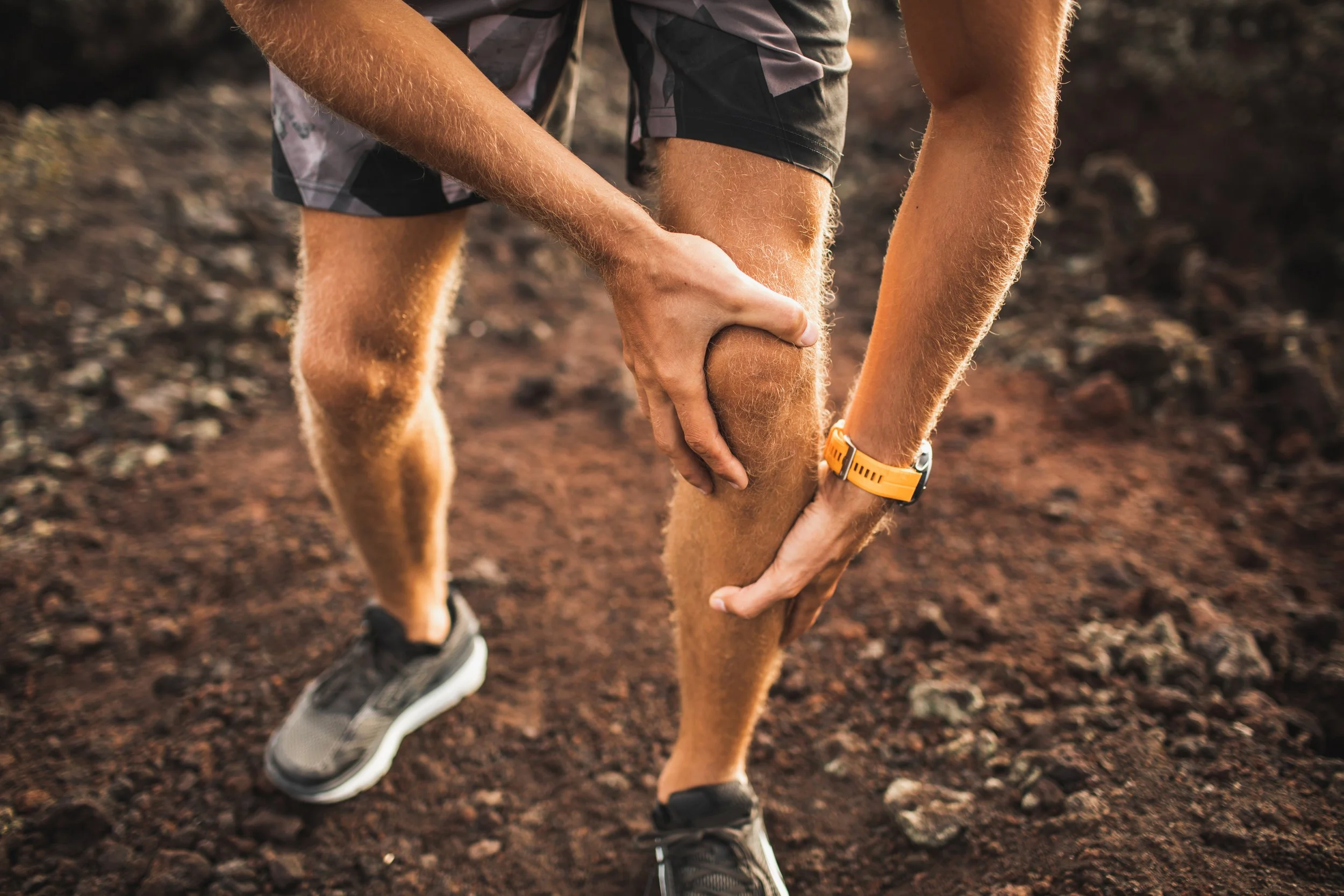 A person wearing gray and black running shoes and gray shorts holds their injured or sore left calf muscle with their hands, outdoors on a dirt trail.