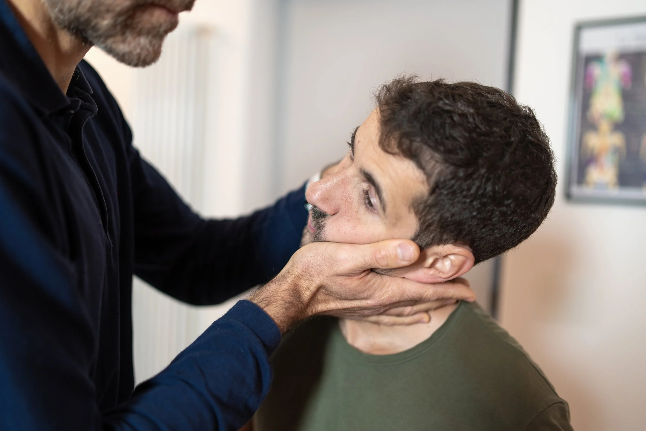 A doctor or chiropractor examines a man's neck and jaw area in a clinical setting.