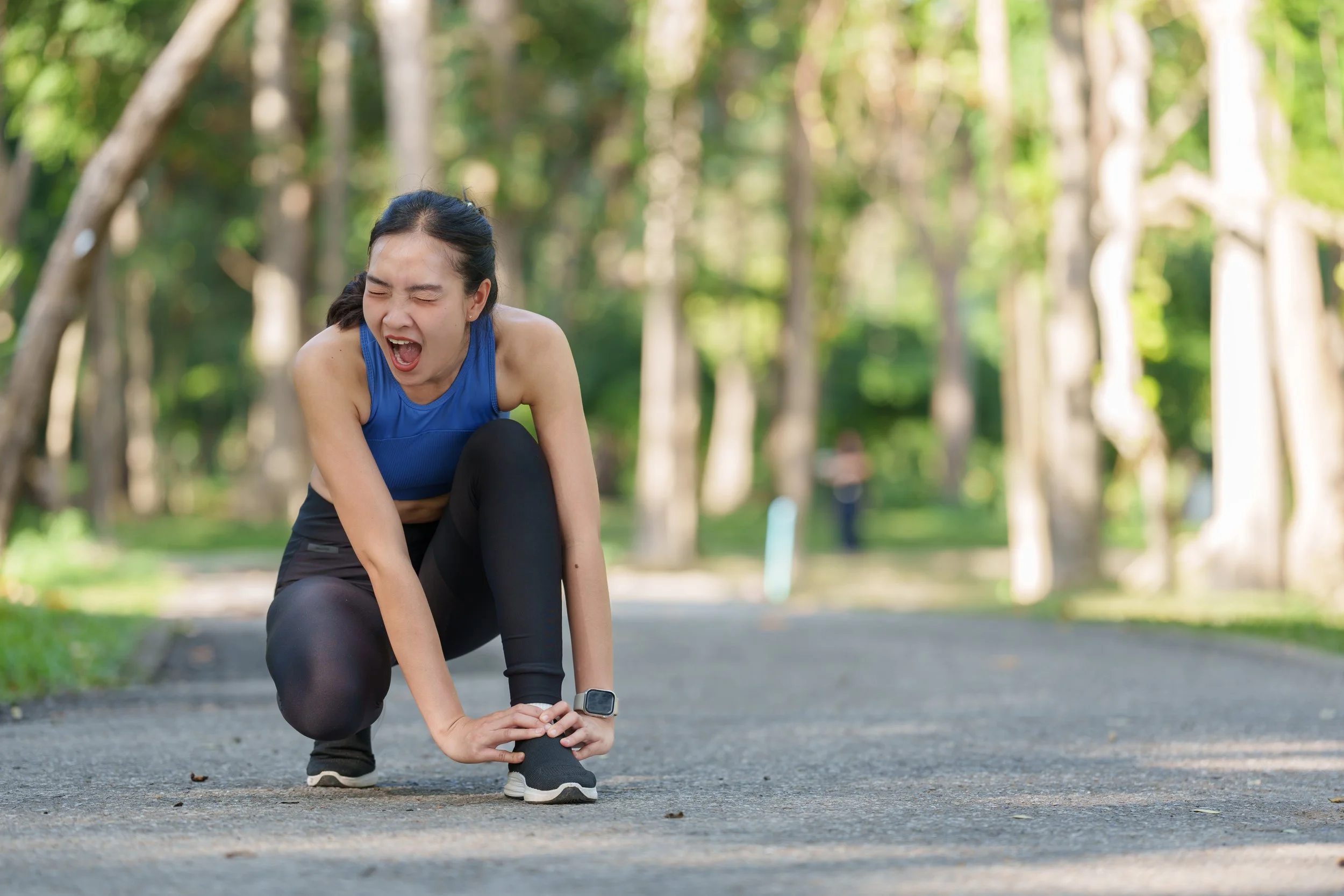 A woman in athletic wear appears to have hurt her ankle while running outdoors in a wooded park and is holding her ankle in pain.