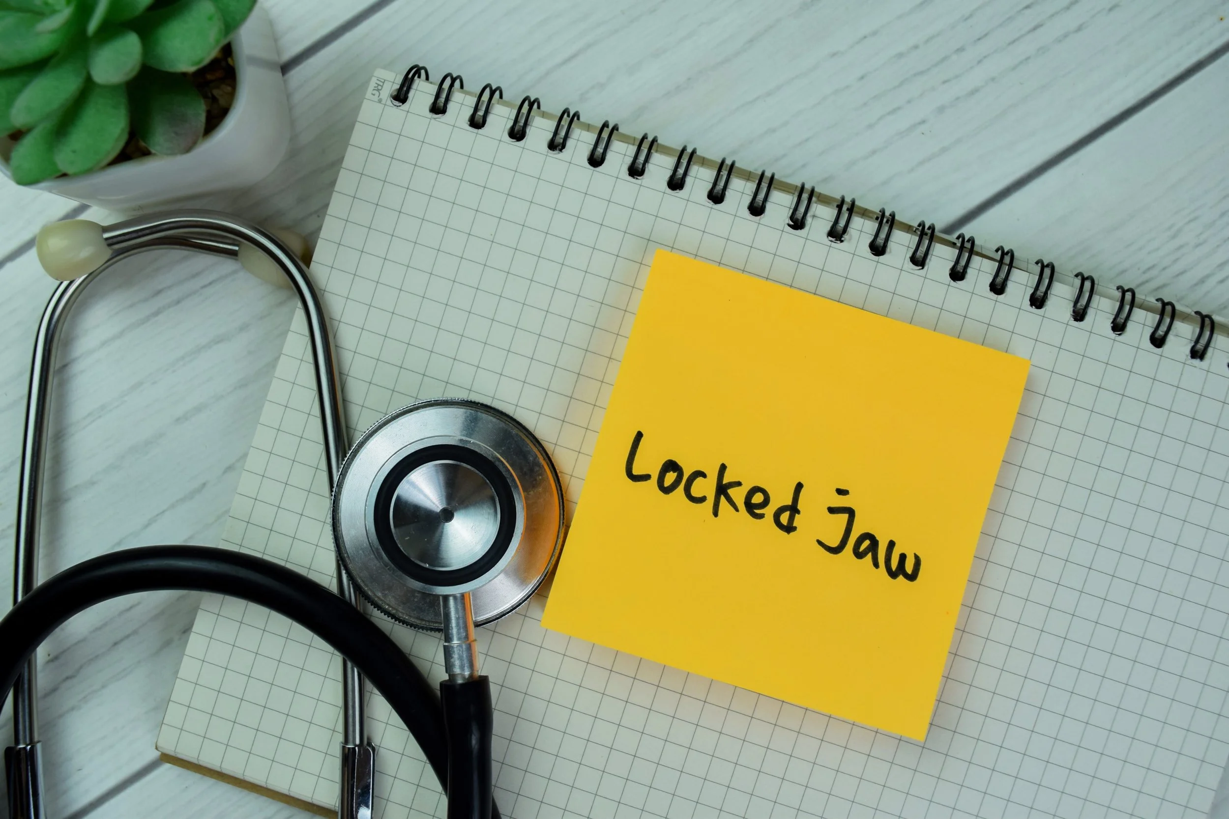 A stethoscope, a yellow sticky note with 'Locked Jaw' written on it, a spiral notebook, and a small potted plant on a white wooden surface.