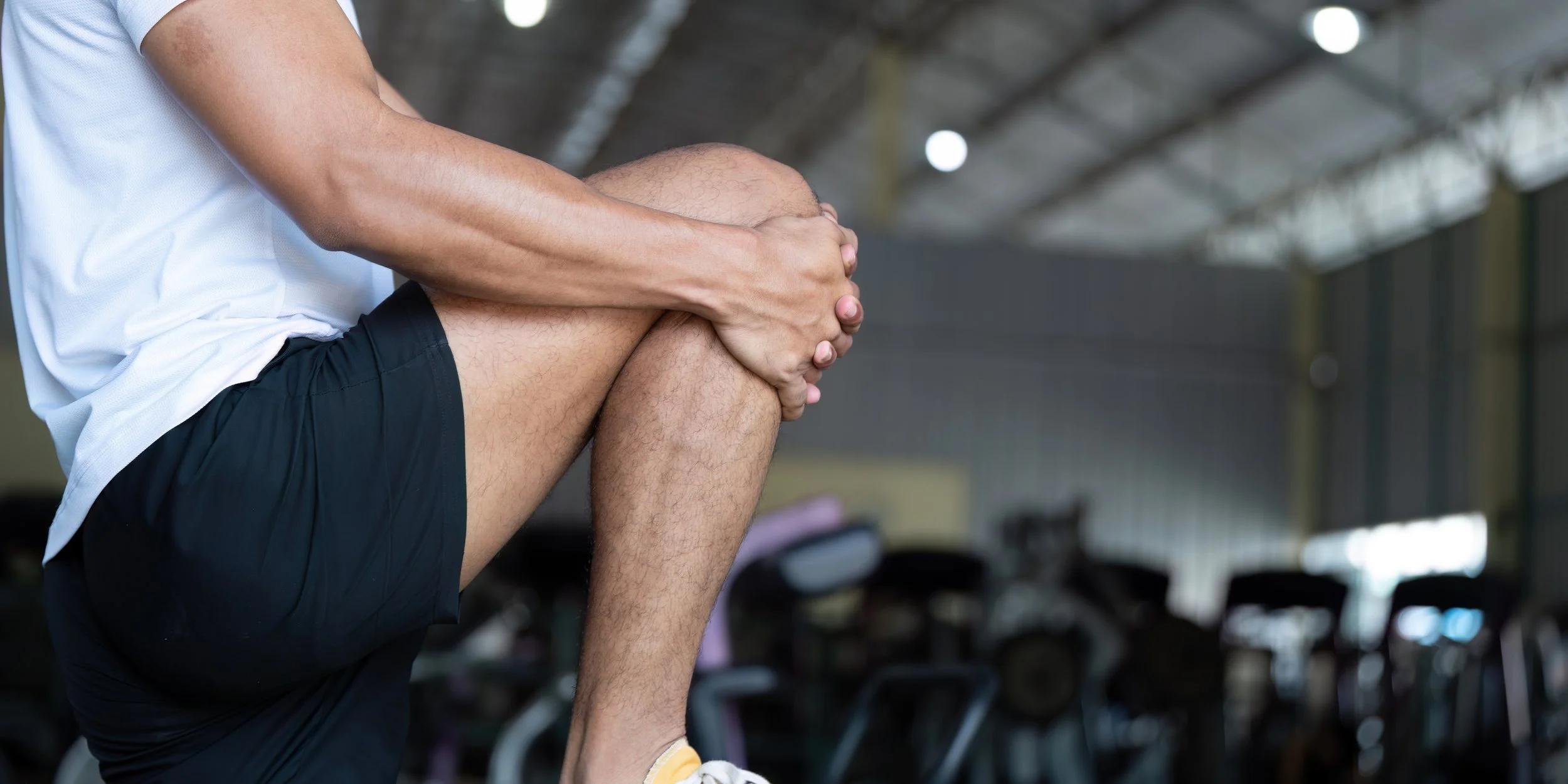 A man in workout clothing holding his knee in discomfort at a gym.