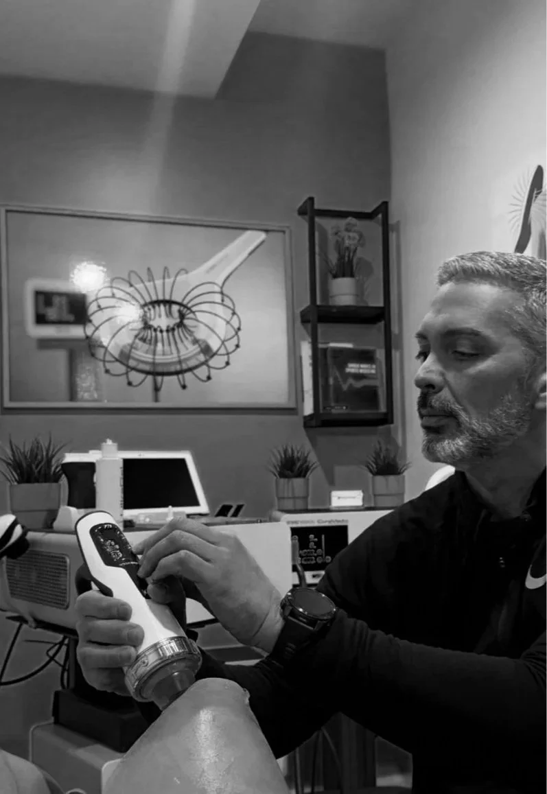 A man with a beard and short hair sitting while holding a massage gun on his leg.