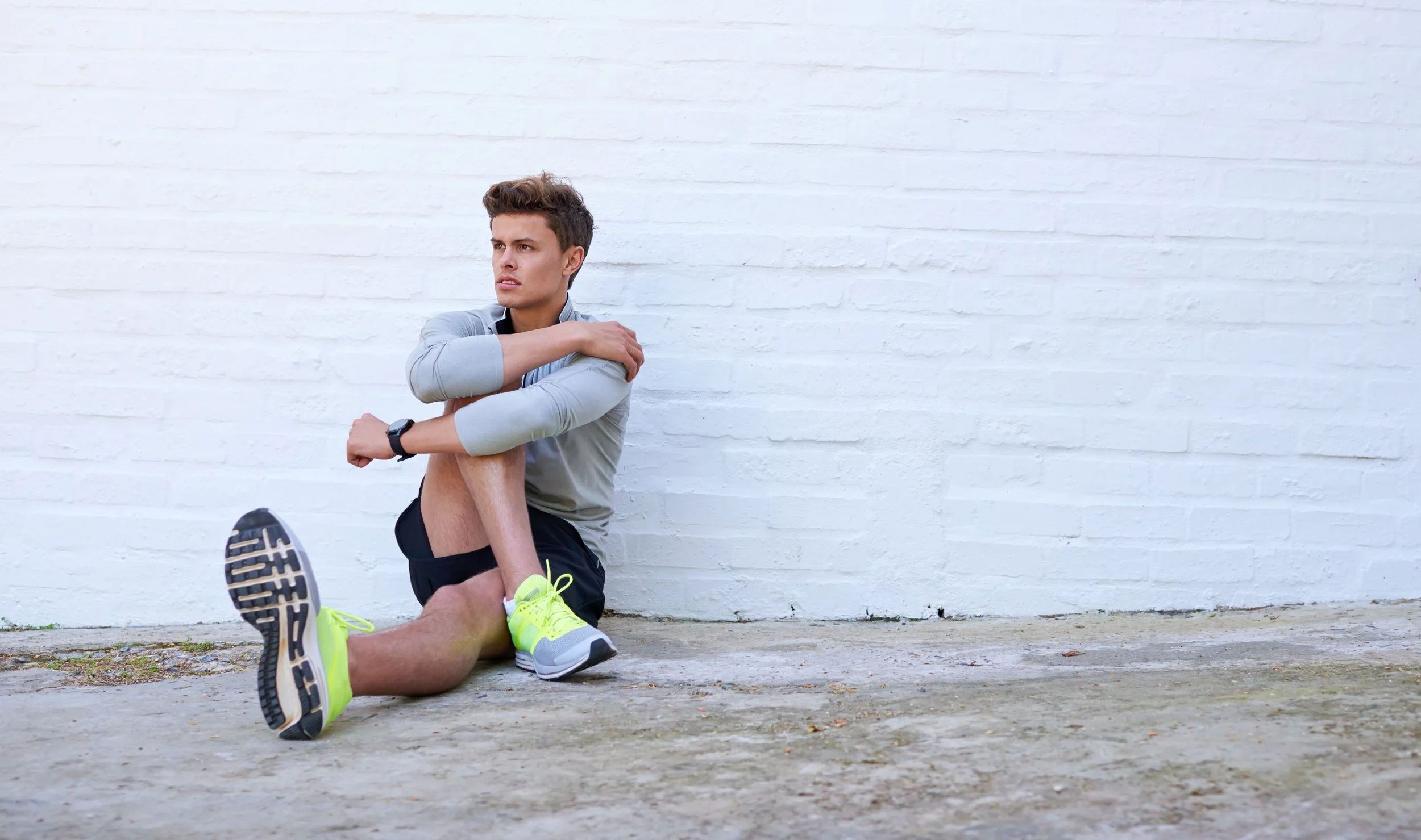 Young man sitting on ground against a white brick wall, looking away with a concerned expression, stretching his leg.