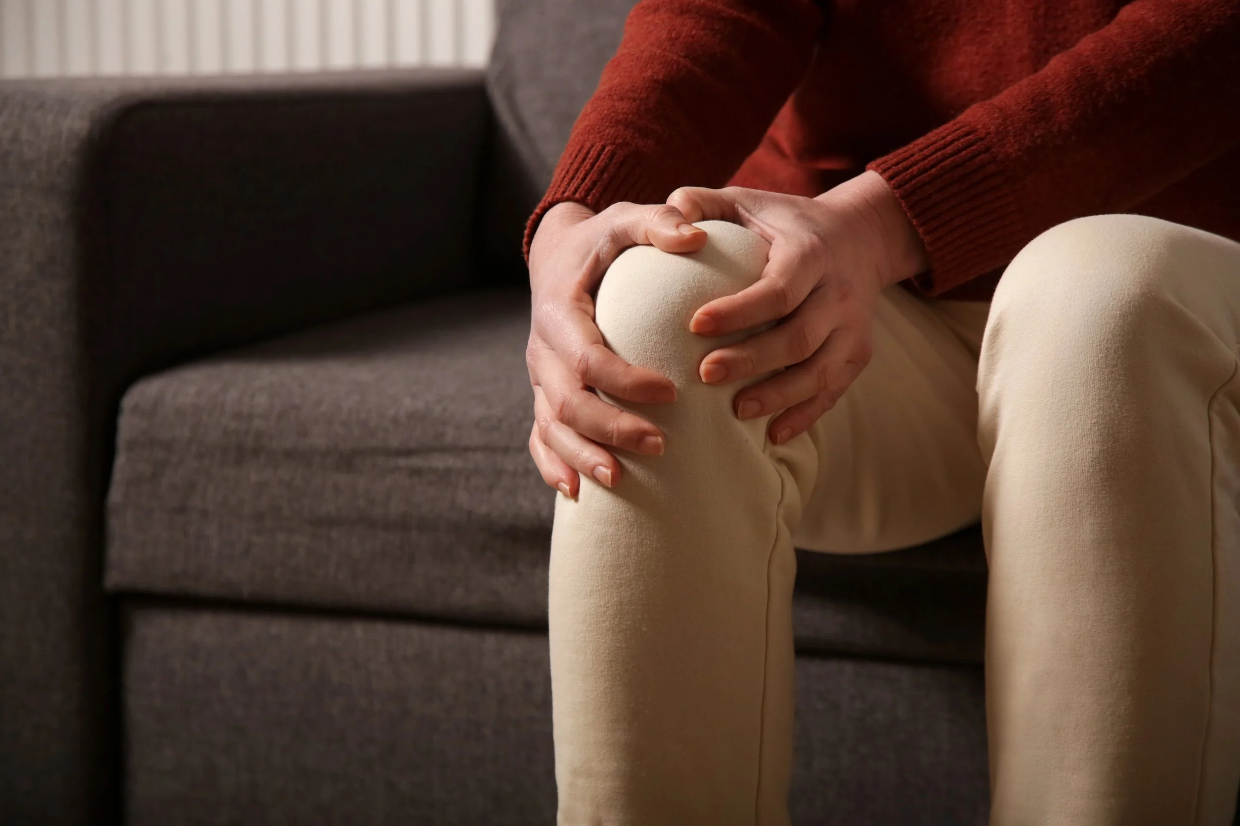Person sitting on a couch holding their knee in pain, suggesting knee injury or discomfort.