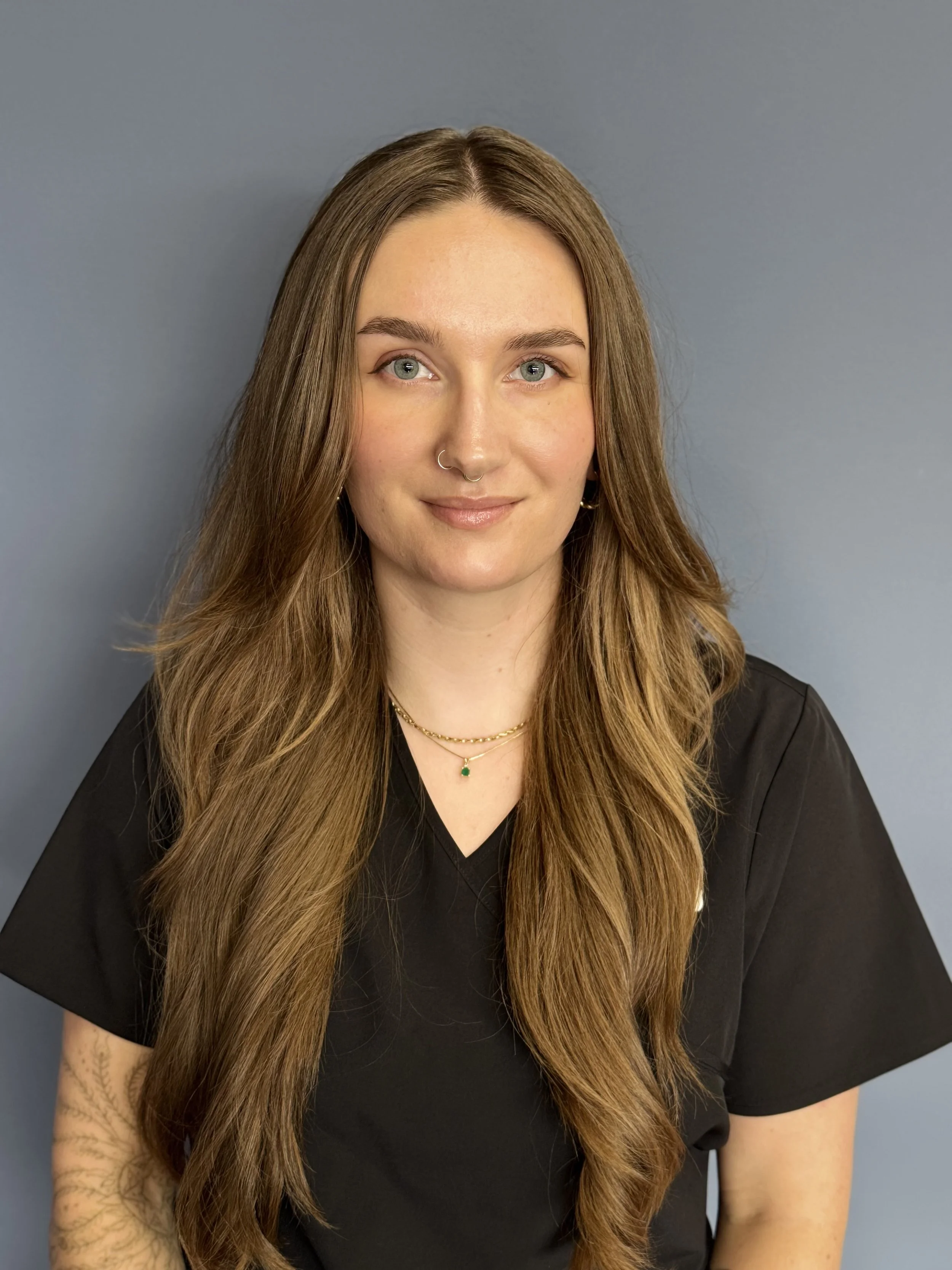 A young woman with long, wavy brown hair, wearing glasses, a teal medical scrub top, and layered gold necklaces, standing against a plain gray background.