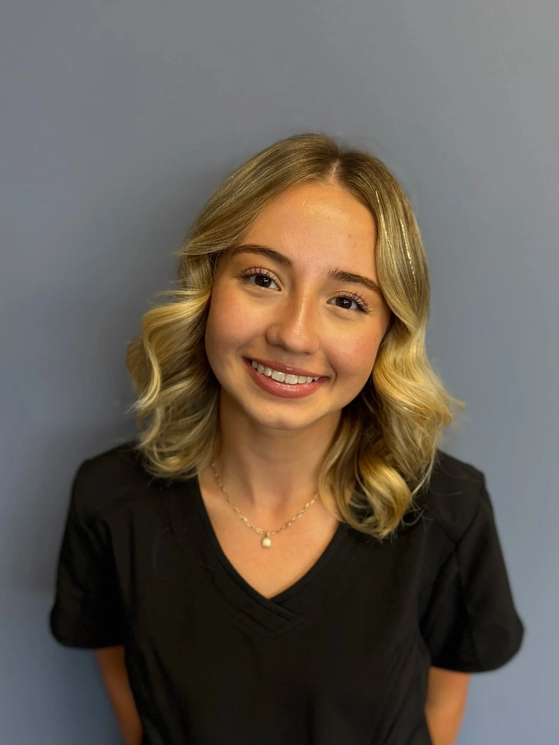A young woman with blonde, wavy hair and a bright smile, wearing a black top and a necklace, standing against a gray background.