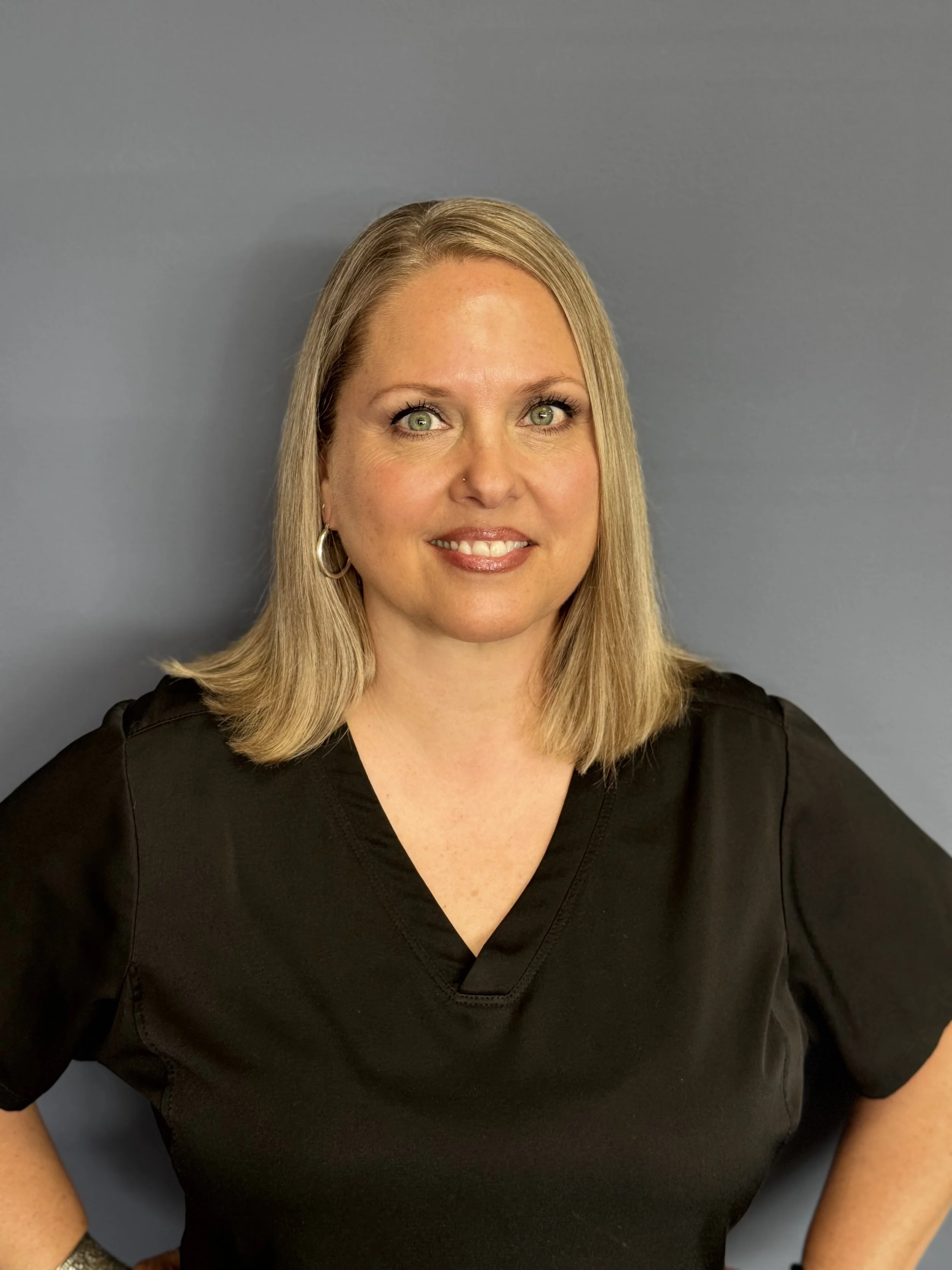 A woman with blonde hair, wearing a navy blue medical scrub top, smiling, standing against a plain gray background.