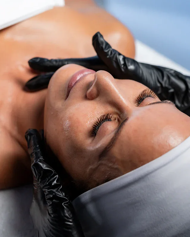 A woman lying on a treatment table during a facial skincare procedure, with a skincare specialist wearing black gloves gently touching her face.