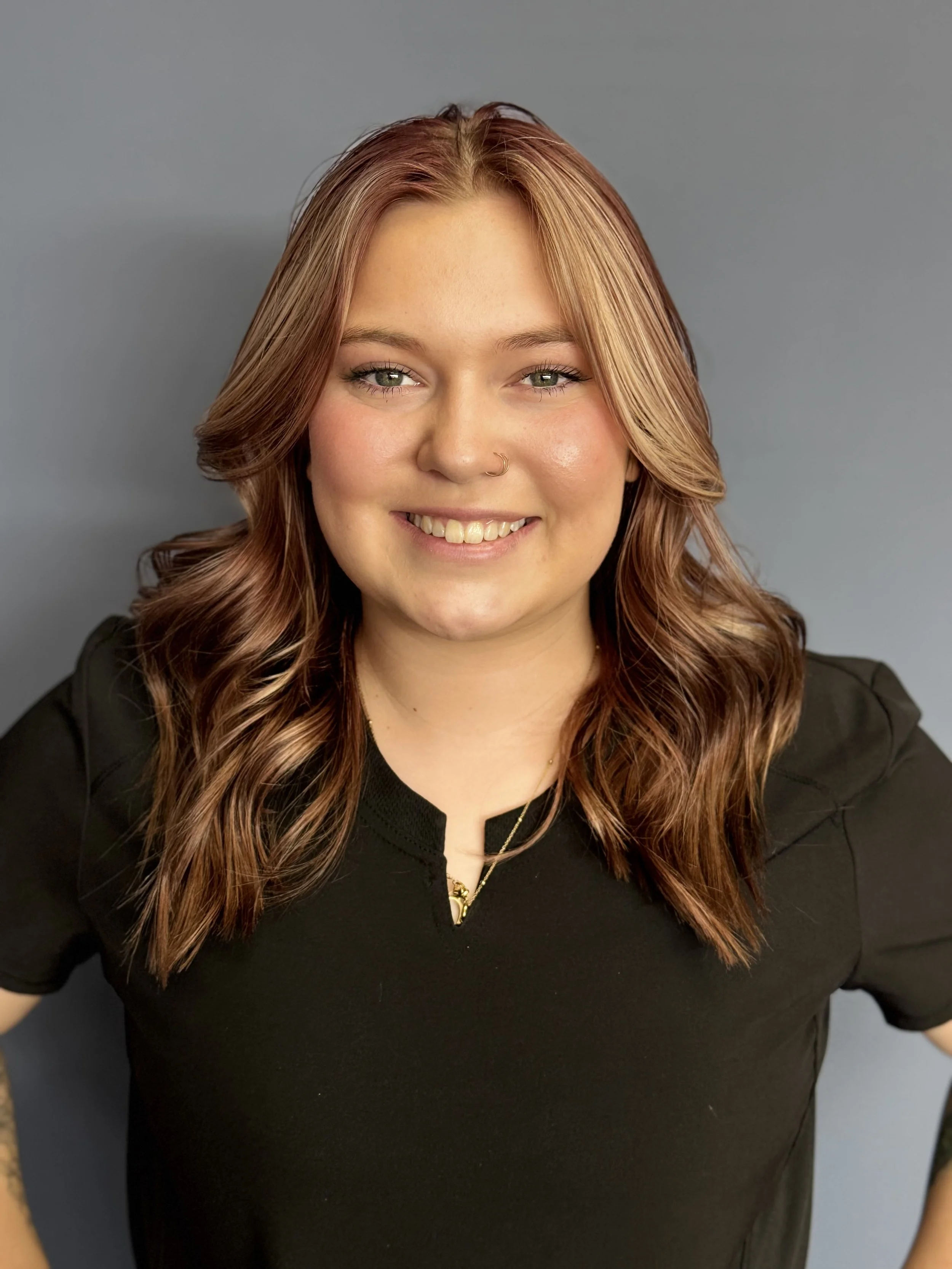 A young woman with blonde hair and tattoos wearing blue scrubs, smiling against a gray background.