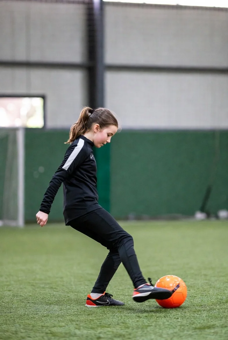 Young girl practicing soccer indoors on artificial turf with an orange soccer ball.