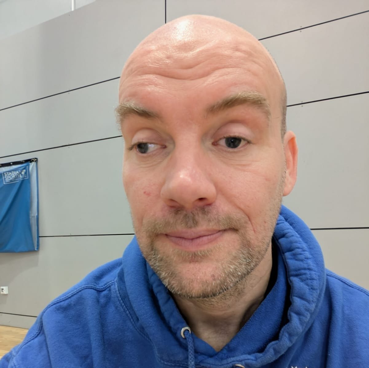 A man with a bald head and light facial hair wearing a blue hoodie, looking slightly to the side, indoors in front of a gray wall with black horizontal lines.