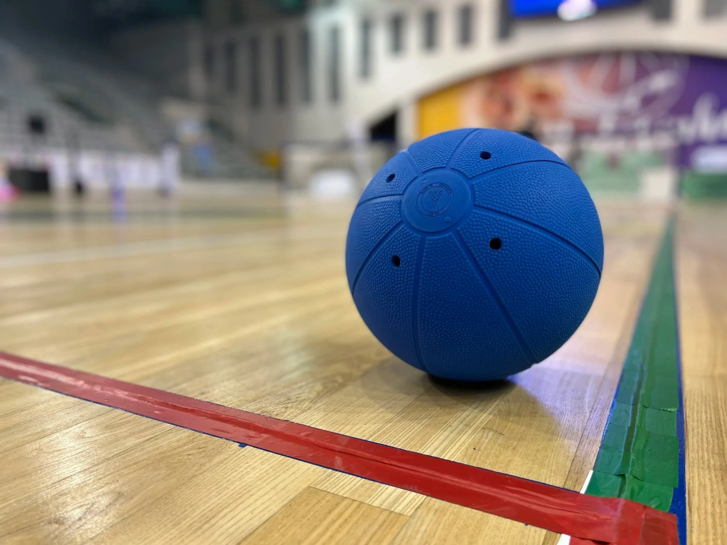 A blue sports ball on a wooden gymnasium court with colored lines and a blurred background of a gymnasium.