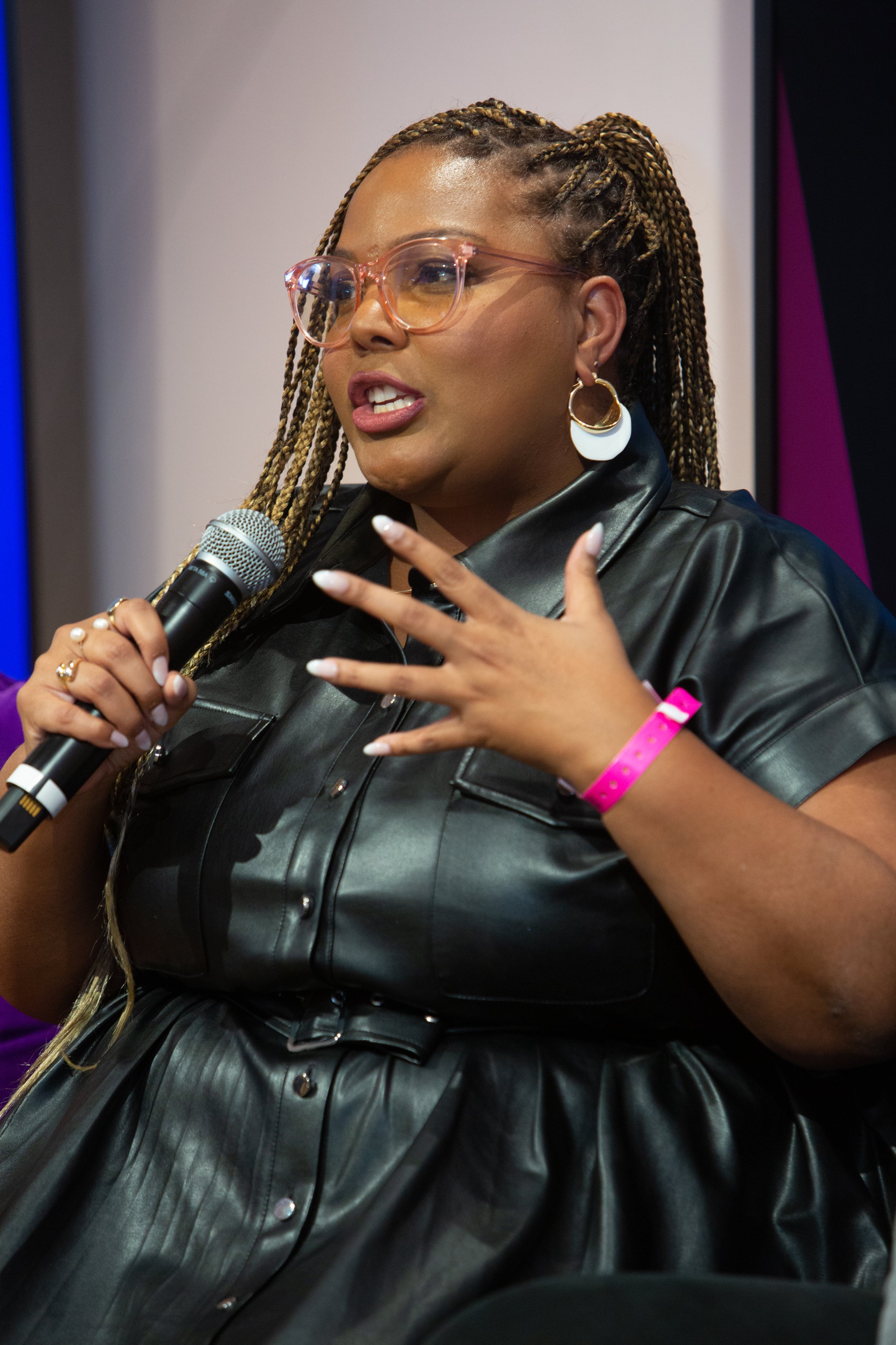 A woman with glasses and braided hair speaking into a microphone during a panel or event.