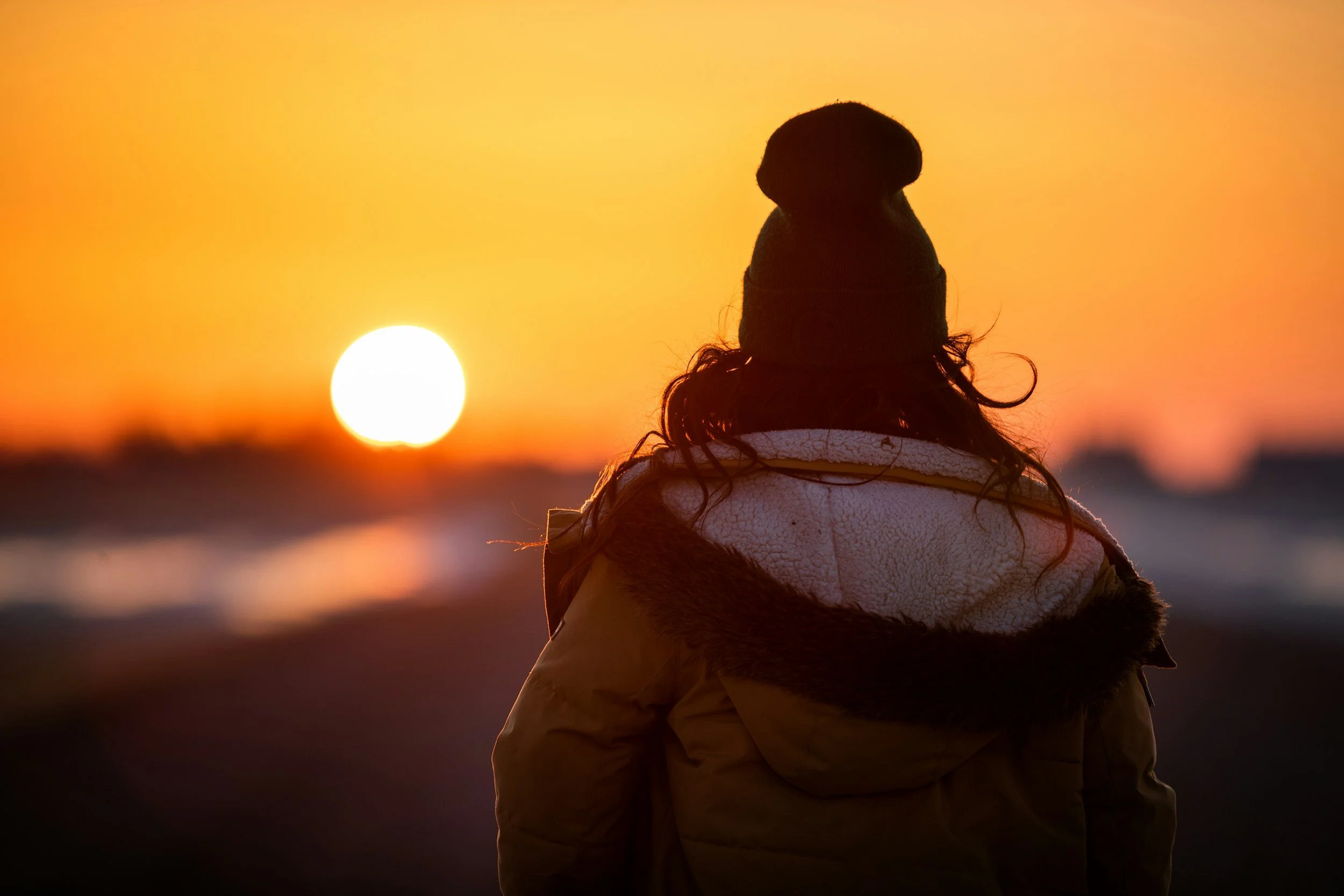 Person in a winter coat and beanie watching a sunset over a landscape.