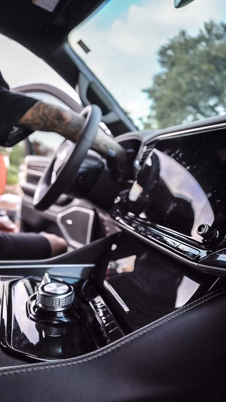 Interior of a modern vehicle, showing the steering wheel, gear shifter, and dashboard with a digital screen. Part of a person's arm with tattoos is visible near the steering wheel.