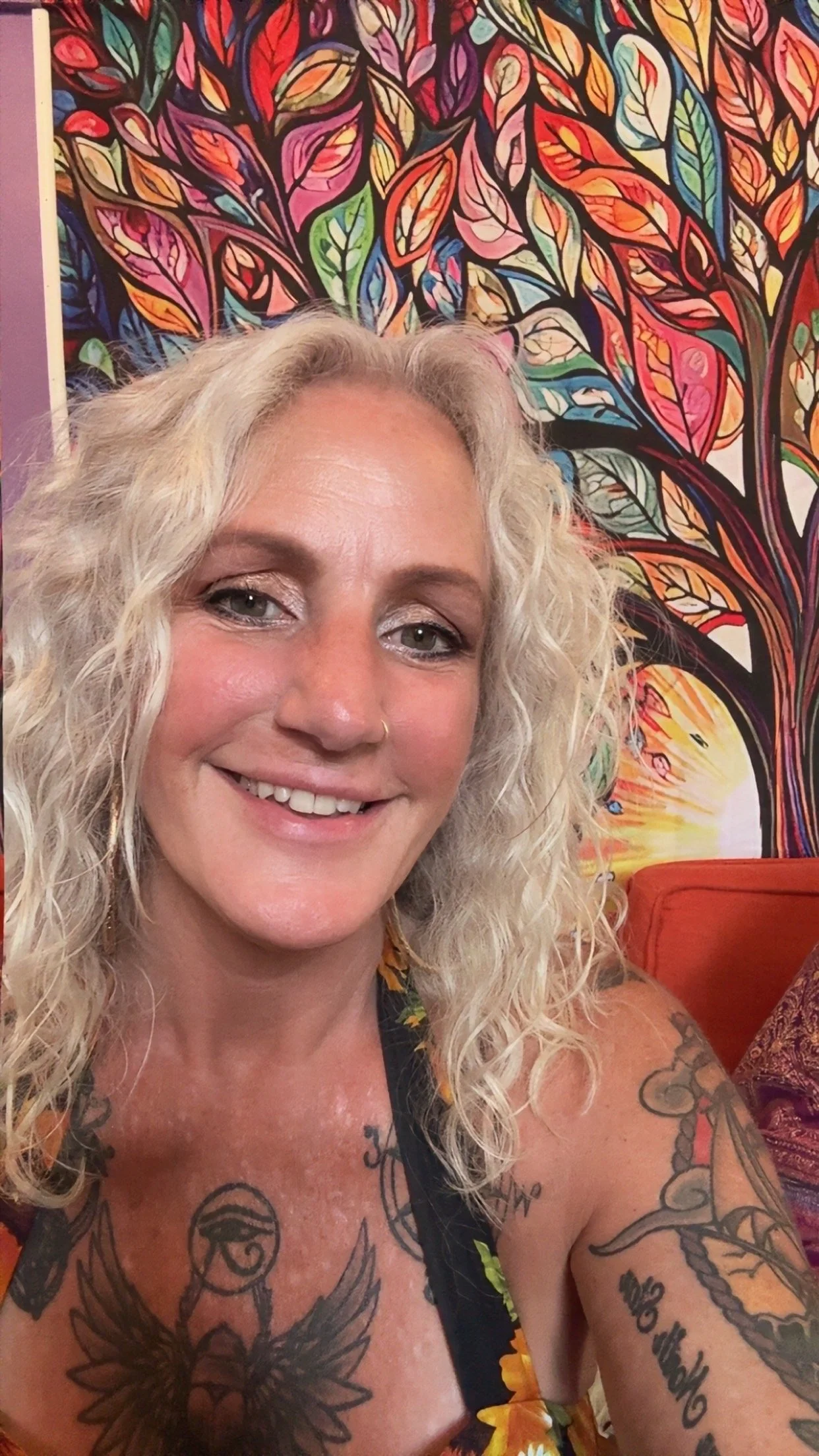 A smiling woman with blonde curly hair and tattoos on her chest and arms, sitting in front of a colorful, abstract tree painting.