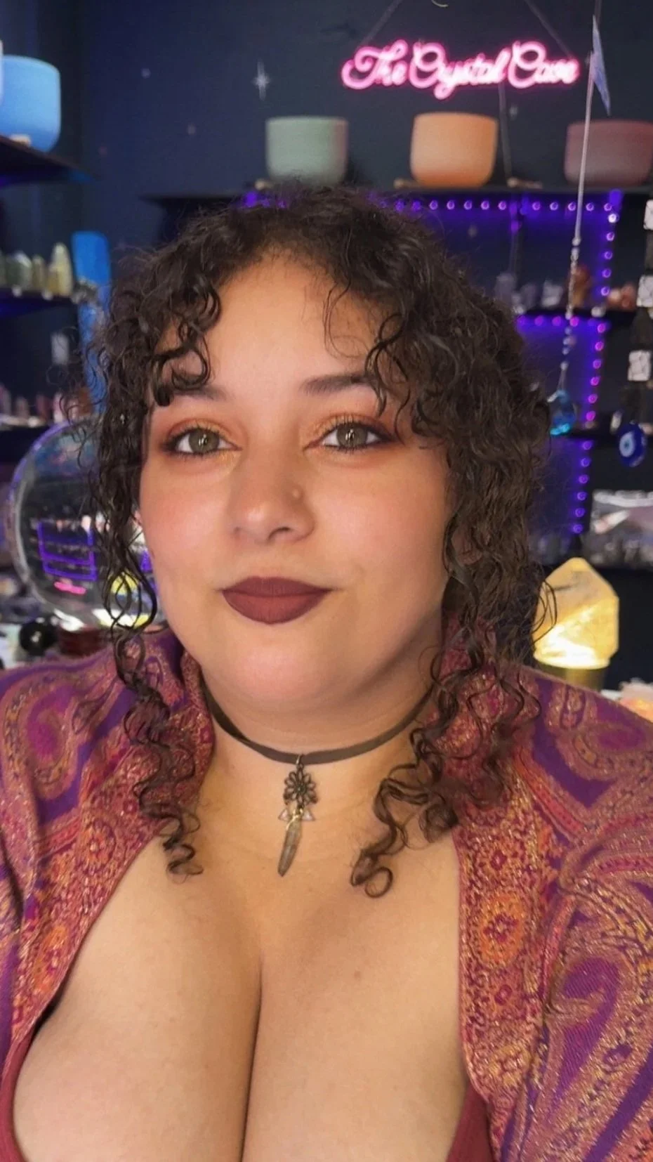 A young woman with curly dark hair and light brown eyes takes a selfie in a room with dark walls decorated with colorful lights and a neon sign that says 'The Crystal Cave.' She is wearing a patterned purple and gold jacket, a black choker with a pendant that appears to be a feather, and has makeup with dark lipstick.