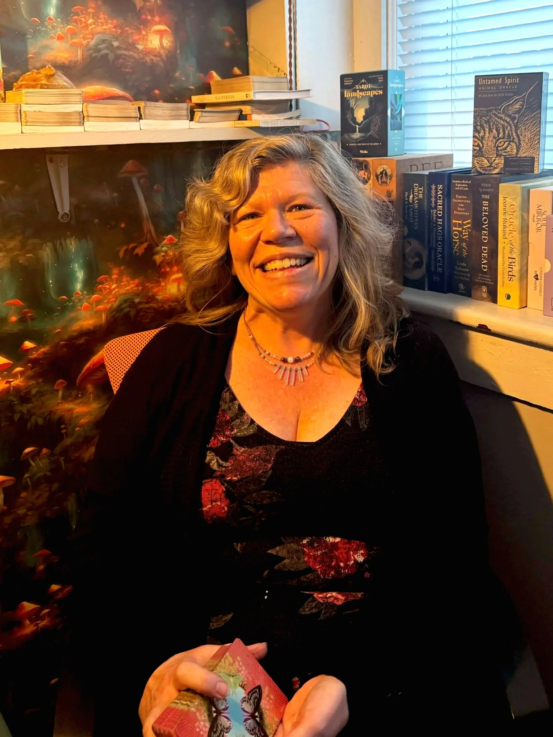 A woman with wavy blonde hair, smiling, holding a deck of colorful butterfly-themed playing cards, sitting in a room decorated with books and artwork, near a window with blinds.
