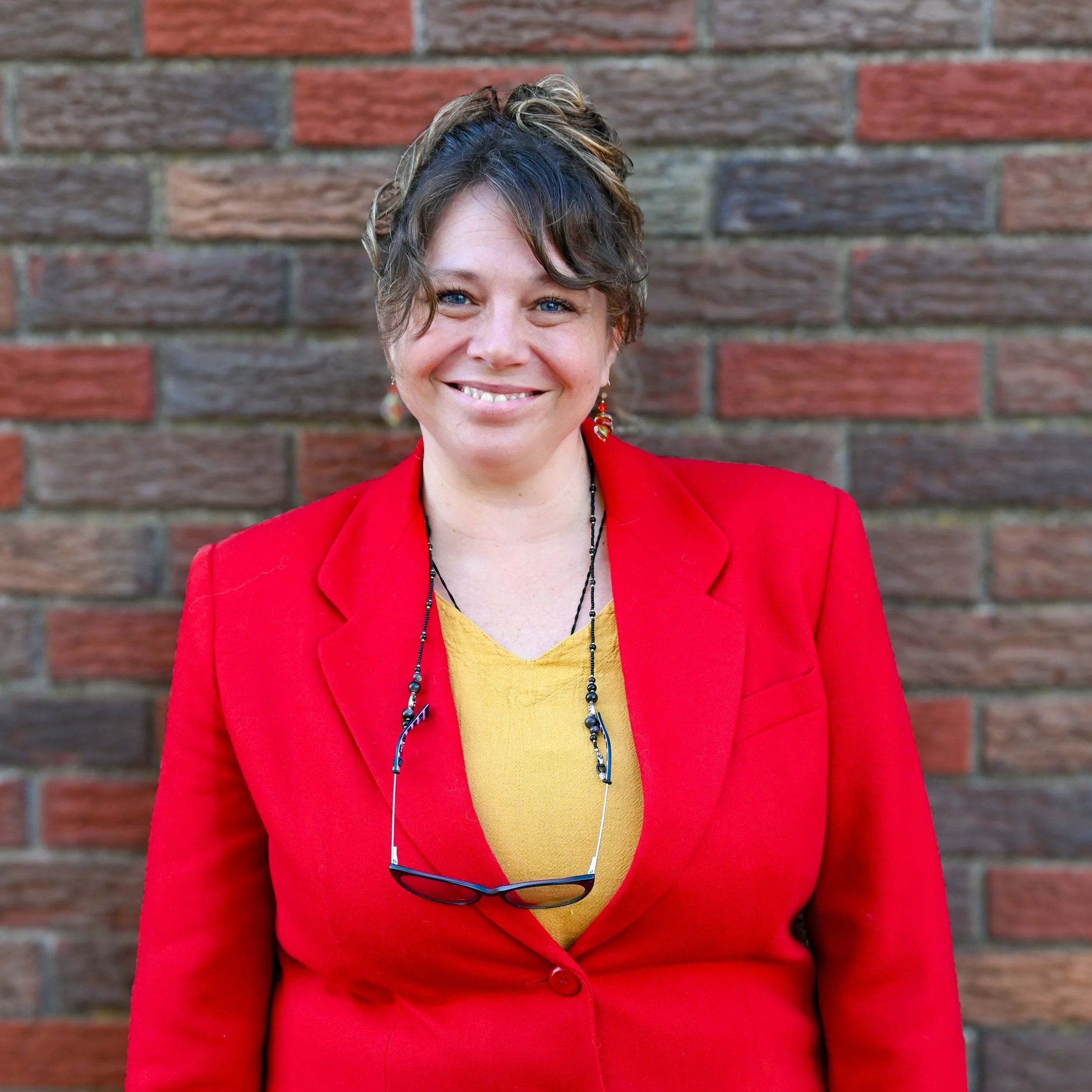 Close-up of a woman smiling, wearing a red blazer, a yellow top, and glasses hanging from her neck, standing in front of a brick wall.