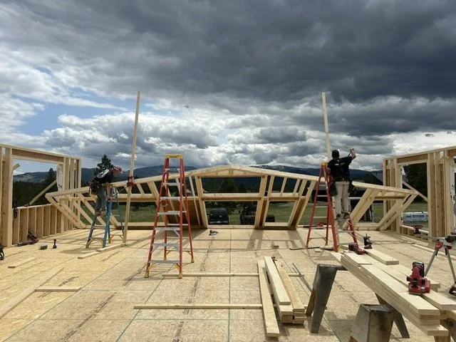 Construction workers building a wooden frame for a house with a scenic mountain view and cloudy sky in the background.