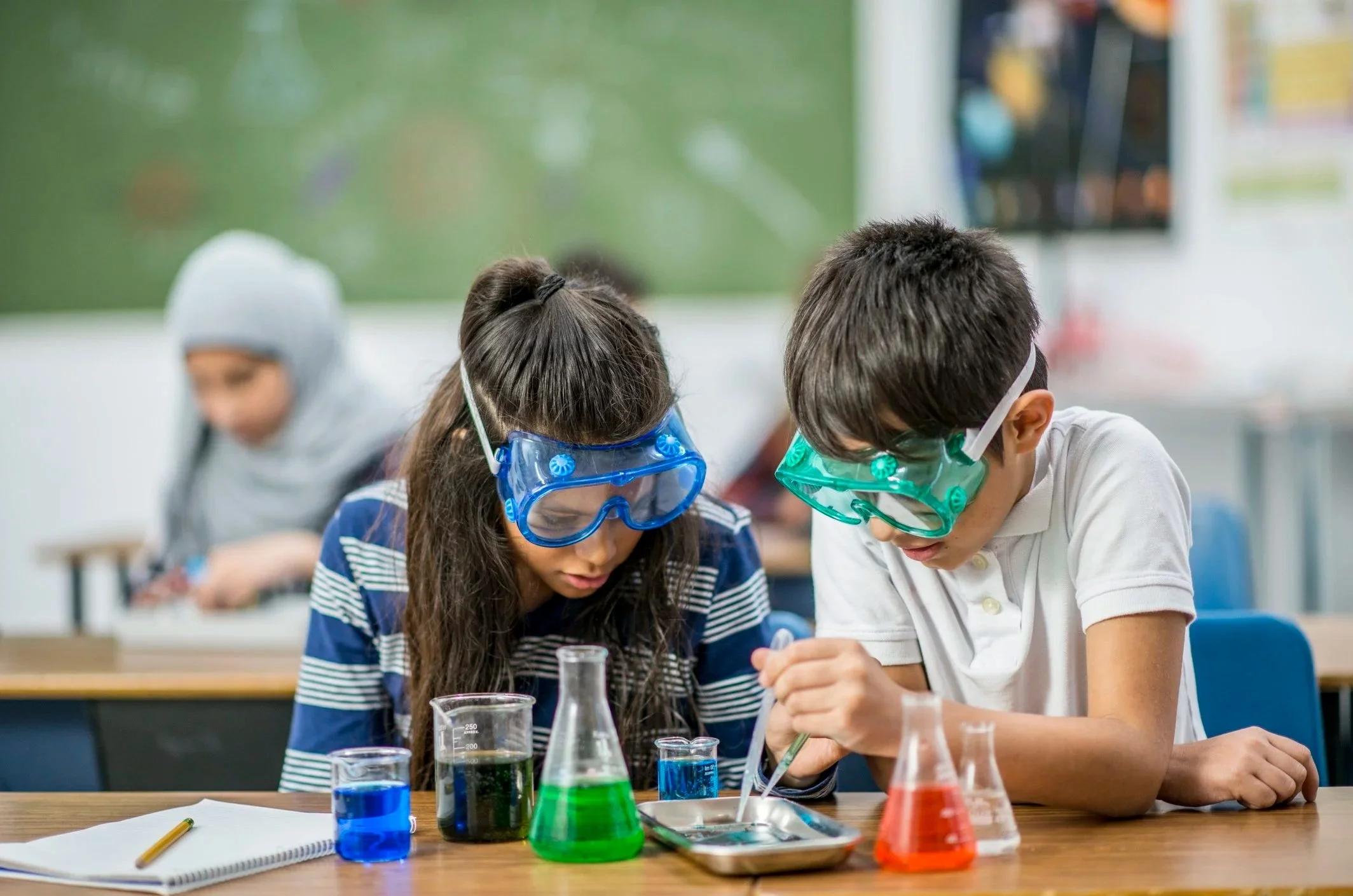Two children with safety goggles conducting a science experiment in a classroom, with a girl in a blue striped shirt and a boy in a white shirt, surrounded by beakers filled with colorful liquids.