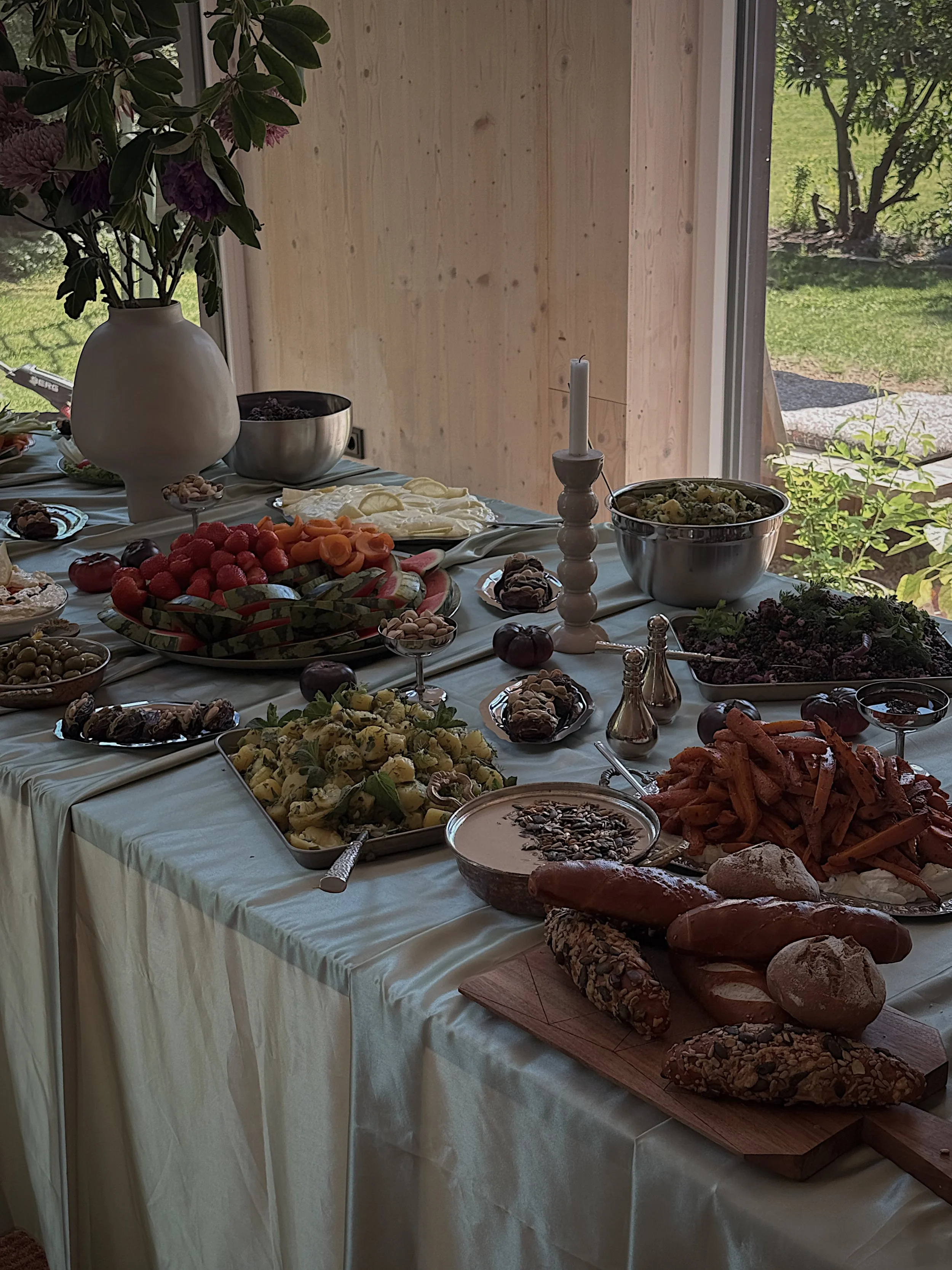 Ein gedeckter Tisch mit verschiedenen Speisen, darunter Obst, Kekse, Brot, ein Salat und andere Gerichte, vor einem Fenster mit Blick ins Grüne.
