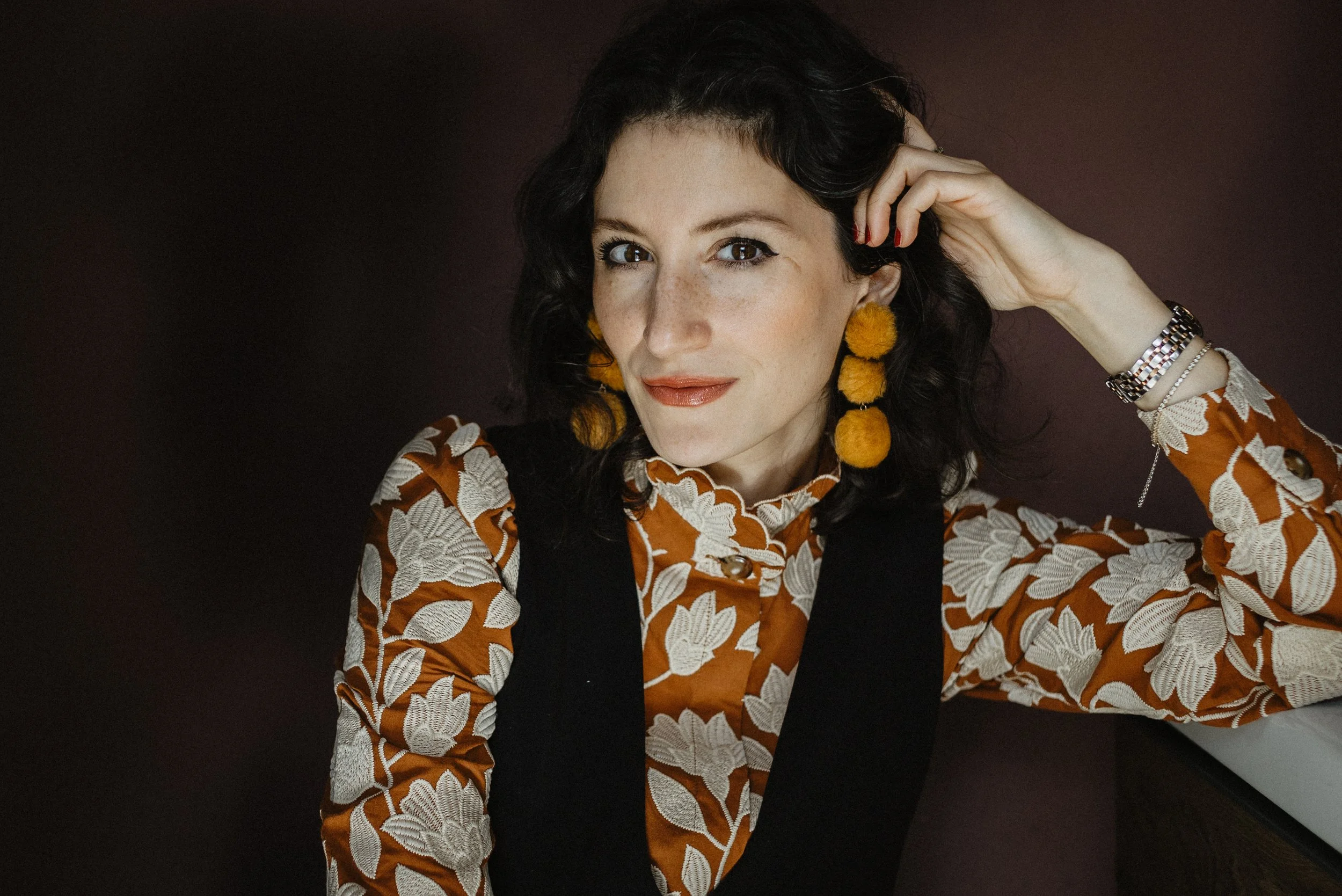 Eine Frau mit schwarzen, lockigen Haaren trägt große, gelbe, pelzartige Ohrringe, einen braun-orangen Pullover mit weißen Blättern und eine schwarze Weste, sowie Armbänder und eine Uhr. Sie sitzt auf einem dunklen Hintergrund und schaut in die Kamera.