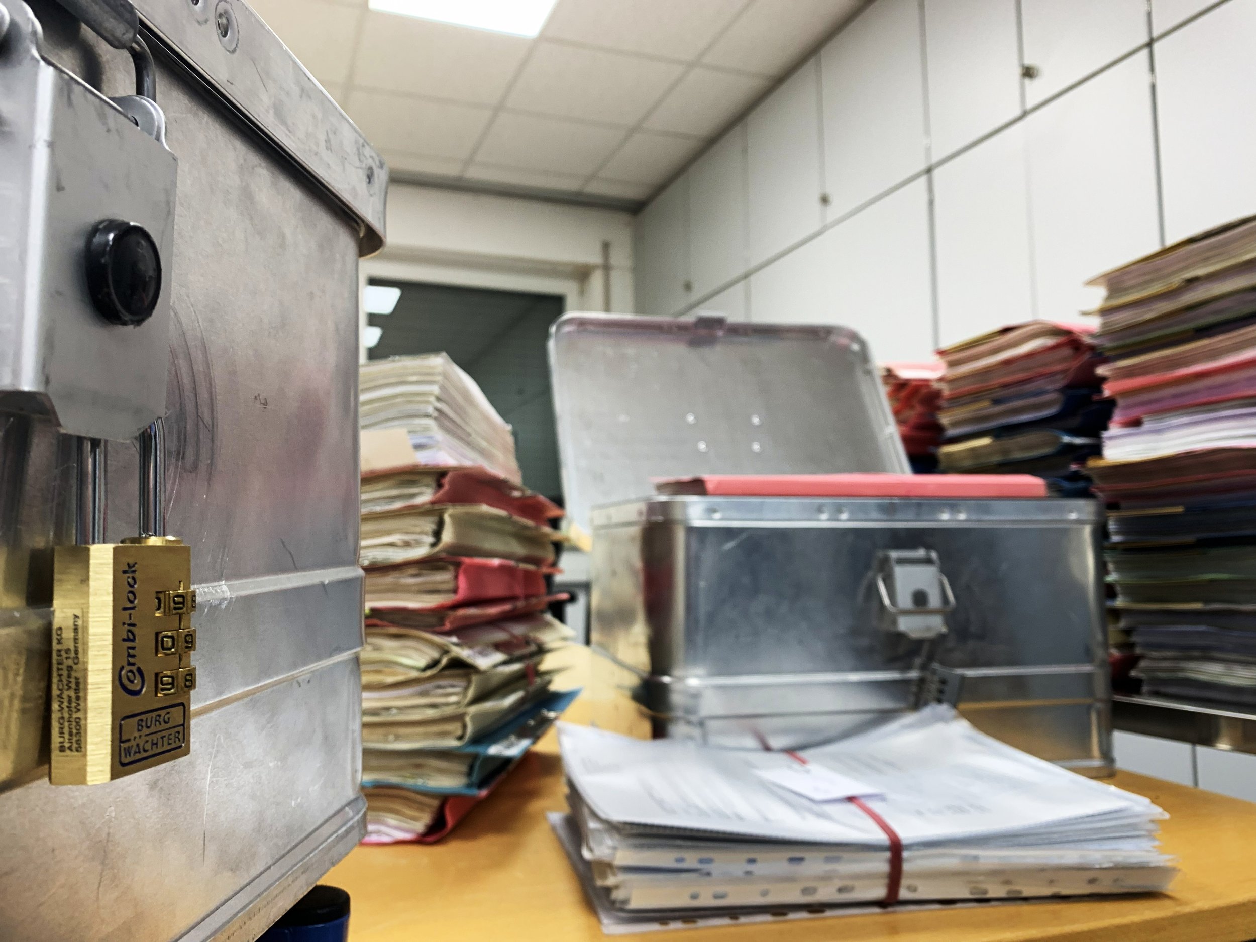 Auf einem Tisch stehen Aktenordner, Papiere und ein datenschutzkonforme Transportbox aus Aluminium mit einem Schloss. Im Hintergrund sind weitere Aktenordner gestapelt und eine Wand mit weißen Fliesen sichtbar.
