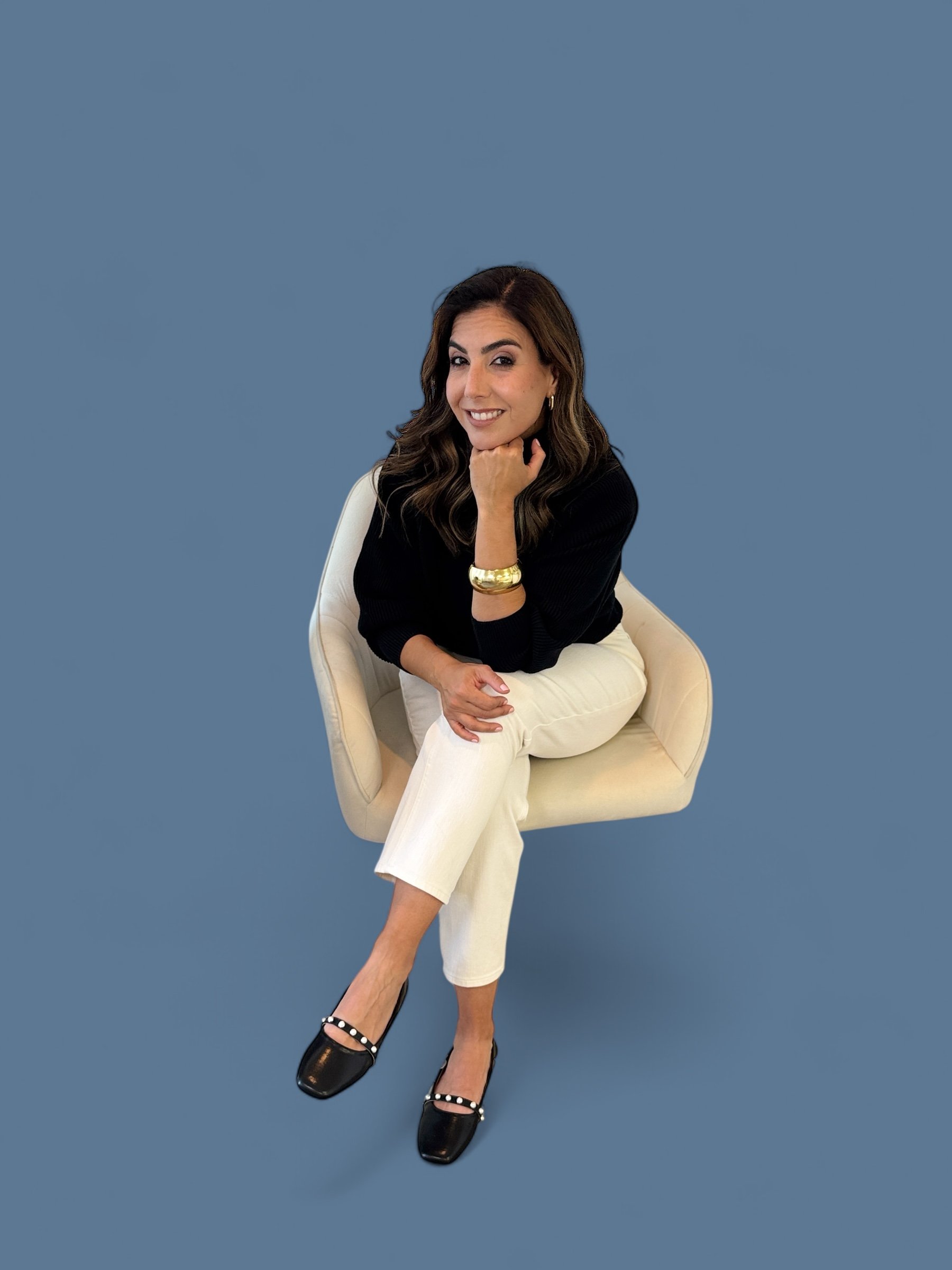 A woman with dark hair and makeup, sitting on a beige chair against a plain blue background. She is smiling, wearing a black top, white pants, black shoes with stud accents, a gold bracelet, and gold hoop earrings. She rests her chin on her hand while looking at the camera.