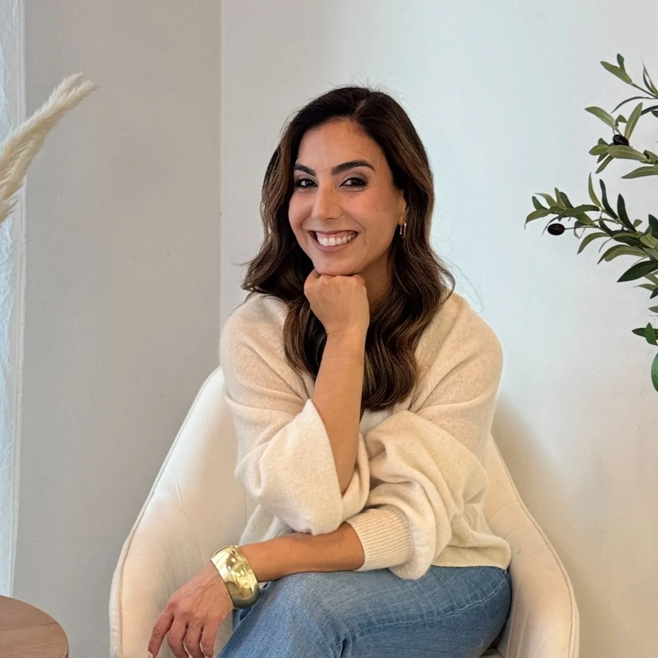 A woman with shoulder-length brown hair, wearing a cream sweater and blue jeans, sitting on a white chair, smiling with her chin resting on her hand, in a well-lit room with a neutral wall and greenery in the background.
