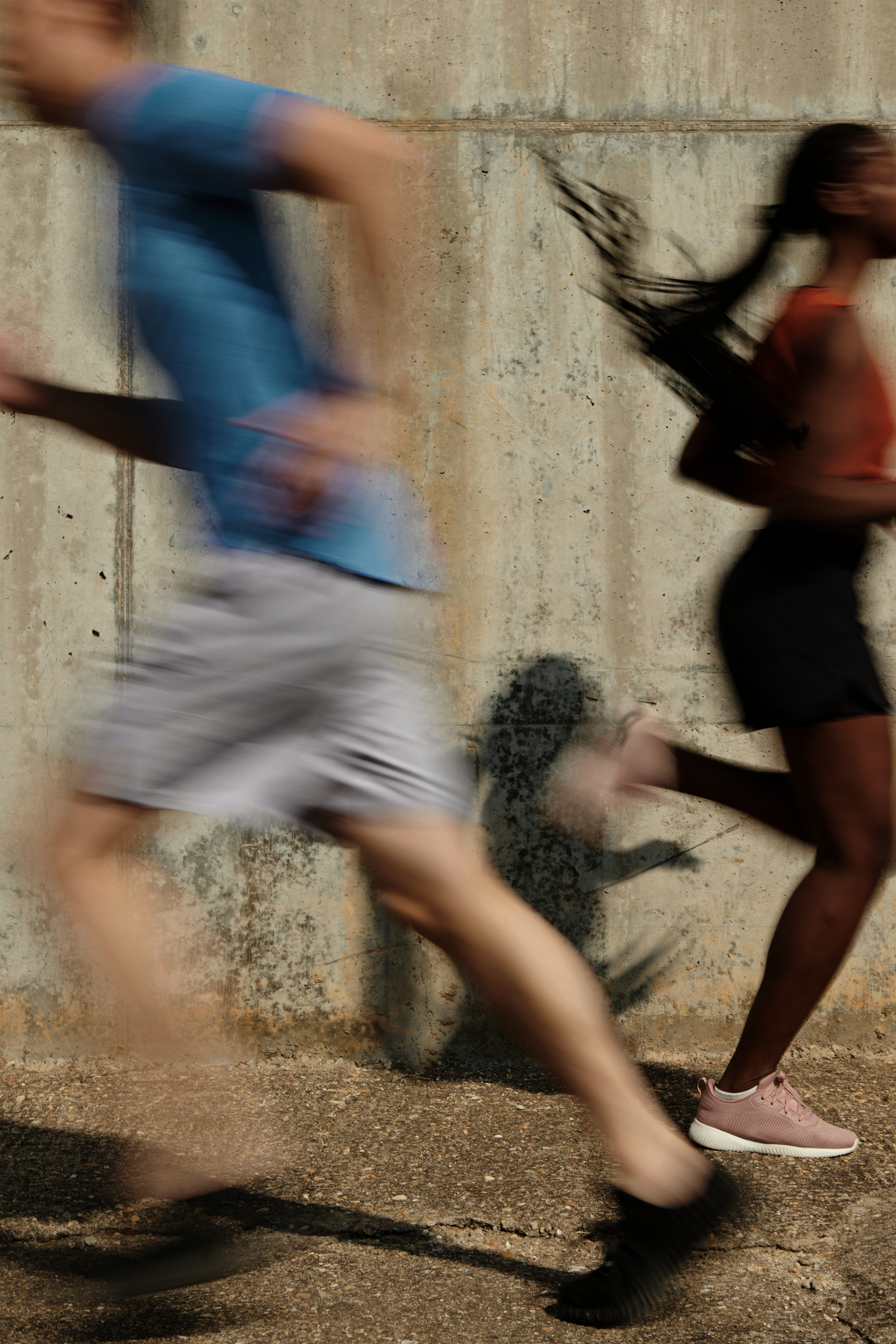 Blurry image of man running in a blue shirt and gray shorts, and a woman running in black shorts and pink sneakers, against a concrete wall.